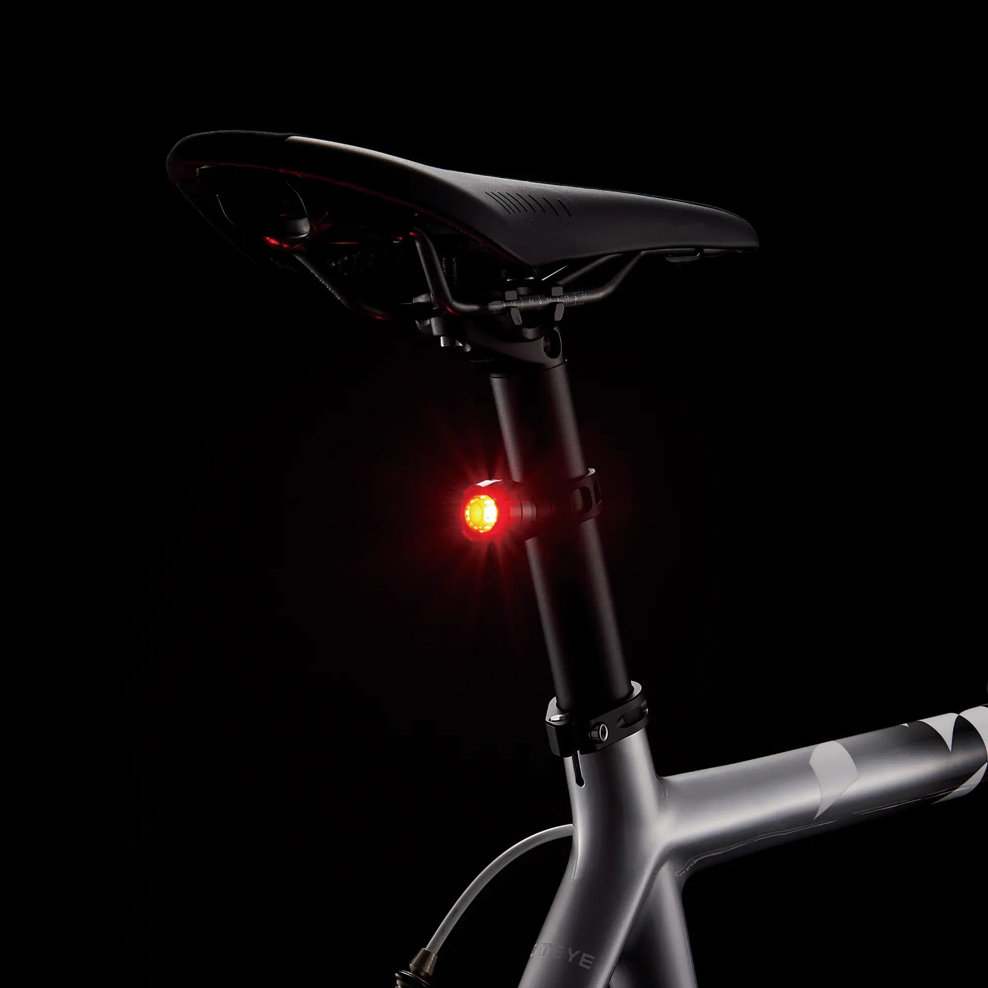 Close-up of a CATEYE VOLT100XC & ORB bicycle seat and seatpost with a small red USB-rechargeable rear light illuminated. The metallic gray frame features sleek, modern lines against a black background.