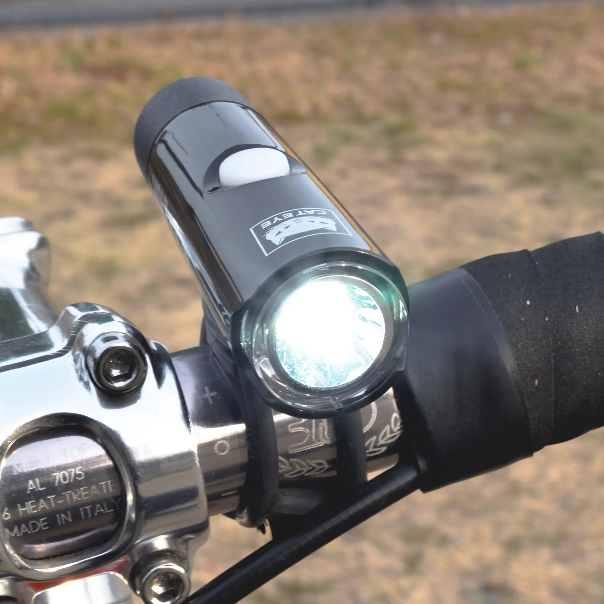 The CATEYE VOLT100XC & ORB, a bright USB rechargeable headlight and taillight combo, is mounted on foam-grip handlebars with metallic parts. The durable aluminum set stands out against a blurred dry grass background.
