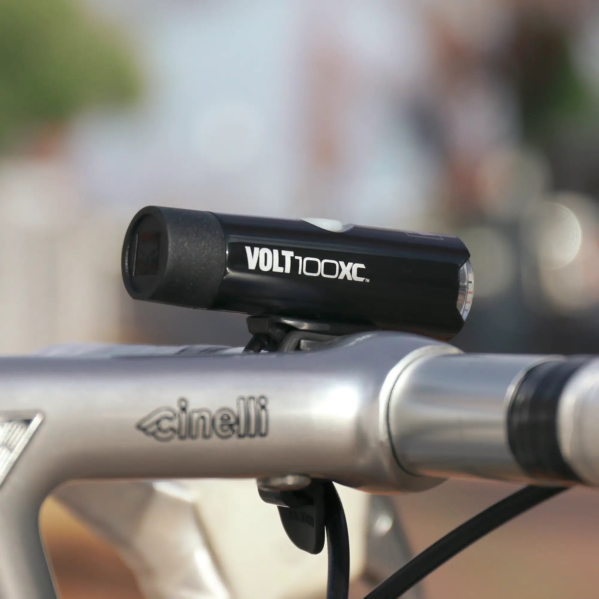 A close-up of a CATEYE VOLT100XC & ORB USB rechargeable headlight mounted on the handlebars of a silver Cinelli bike, set against a blurred outdoor background.