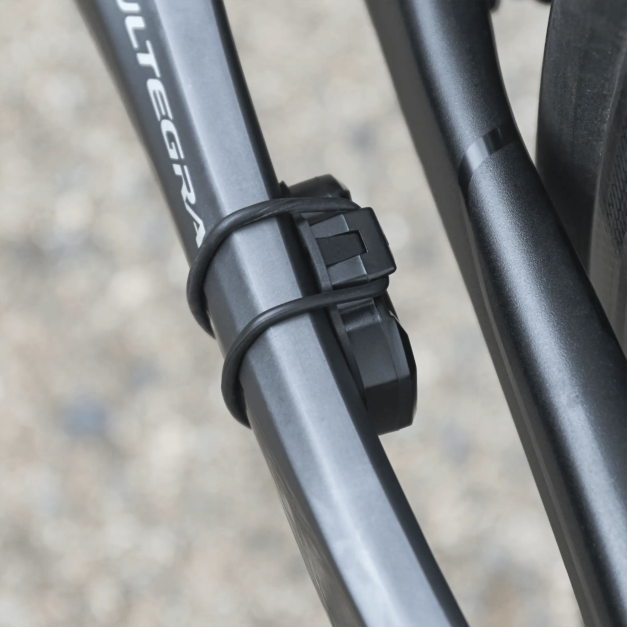 A close-up of a bicycle frame featuring the CATEYE AirGPS sensor secured with zip ties. The ULTEGRA logo is partly visible, while a blurred gravel background emphasizes GPS accuracy for cyclists.