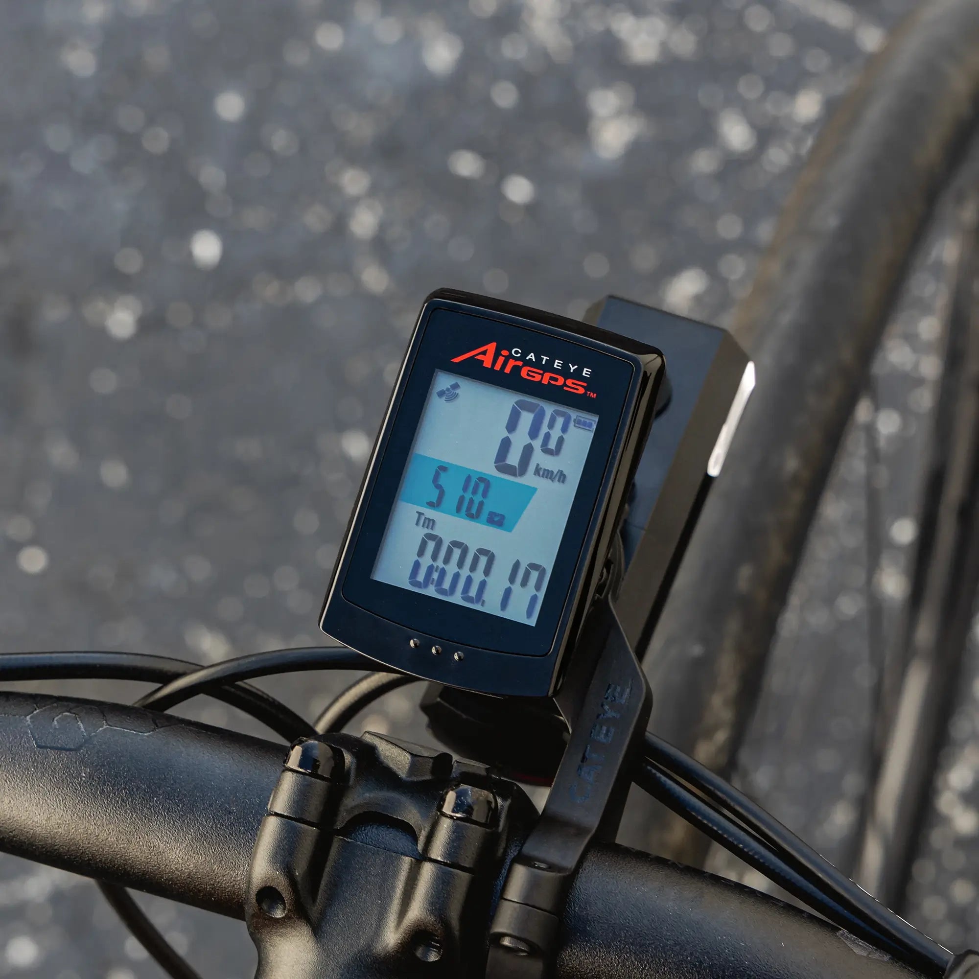 A CATEYE AirGPS bike computer, mounted on the handlebars, displays precise GPS-based speed, distance, and ride time as the bike sits on a paved surface speckled with white dots.