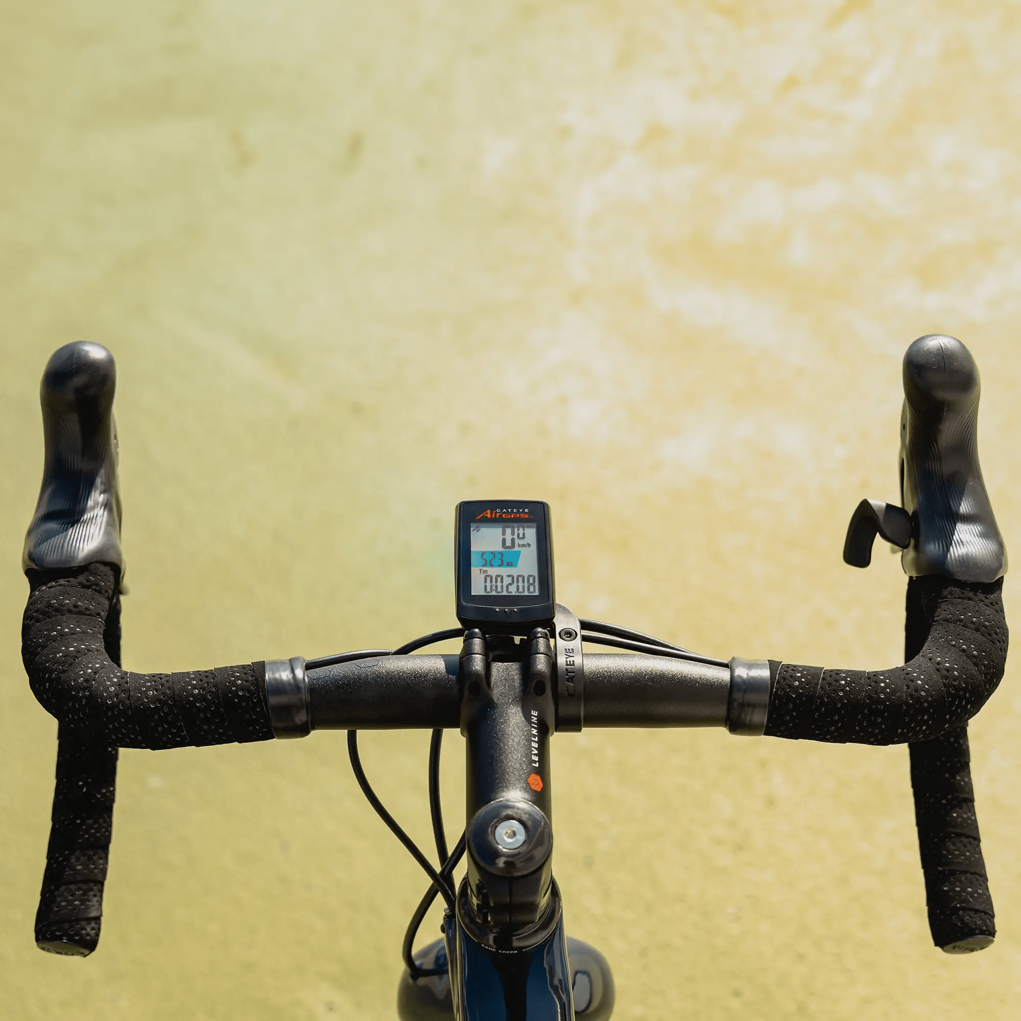 A close-up of bicycle handlebars wrapped in tape, featuring the CATEYE AirGPS cycling computer for enhanced GPS accuracy, set against a light yellow textured background.
