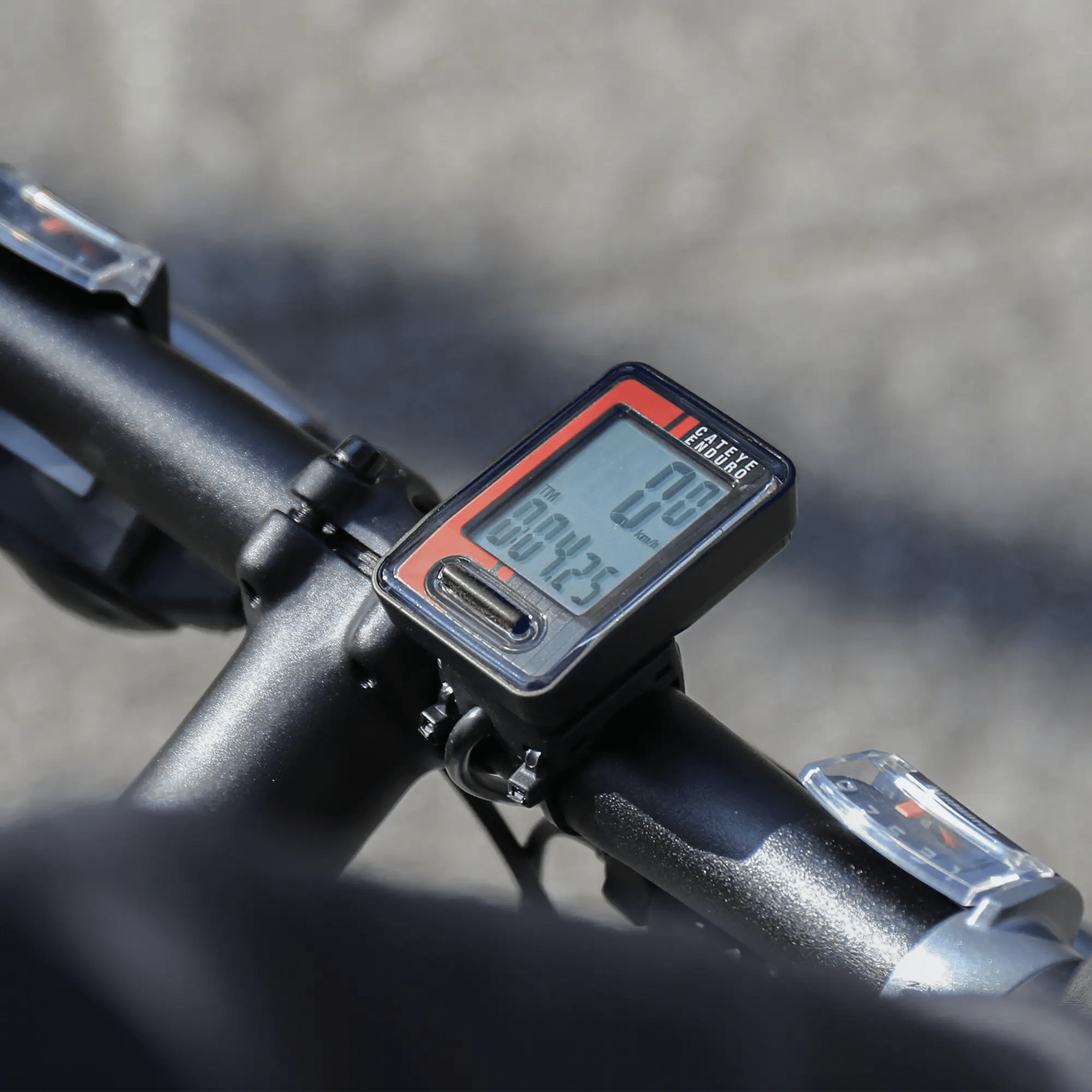 A close-up of a CATEYE Enduro (CPSC) digital bike speedometer with a zip-tie mount attached to handlebars displays tracking data, ideal for adventurous cyclists, against a blurred outdoor background.