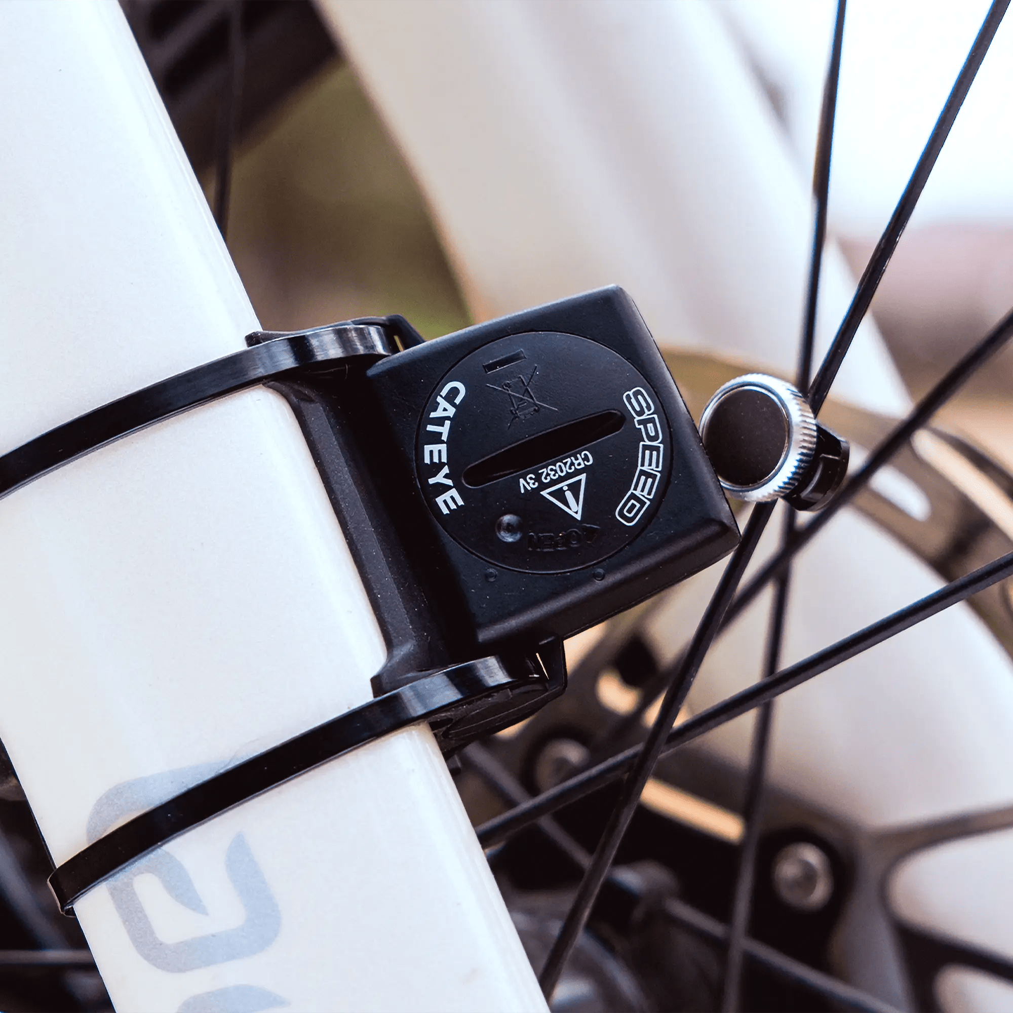 Close-up of a CATEYE Padrone (CPSC) speed sensor secured with zip ties to a bike fork near the wheel spokes; part of the bike frame and the large display are visible in the background.