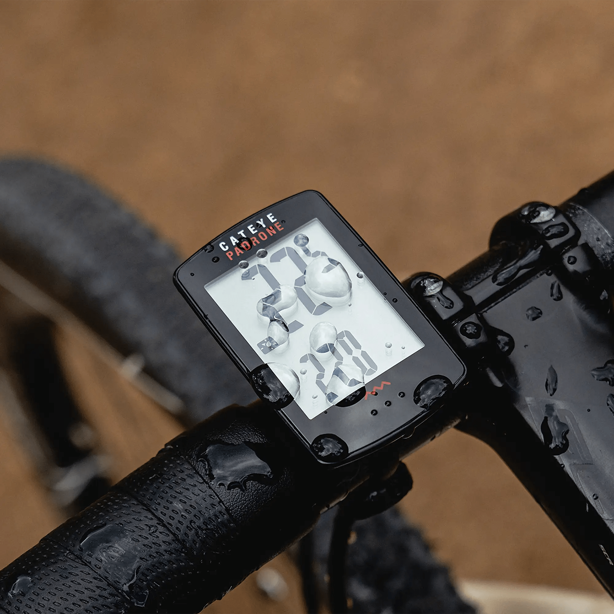 A close-up of a rain-soaked CATEYE Padrone (CPSC) bike computer with a large display, mounted on a bicycle handlebar and showing speed and other metrics. The blurred background shows the bike tire and ground.