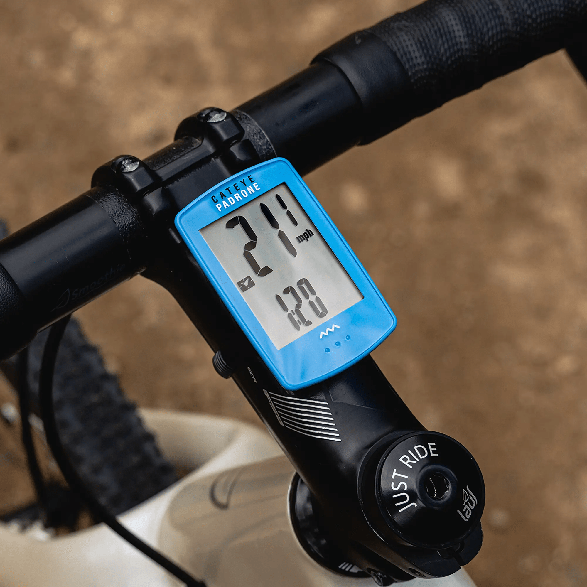 A close-up of a blue CATEYE Padrone (CPSC) with its large display shows 21.7 mph speed and 1.20 odometer, mounted on a bike handlebar with part of the front wheel visible.