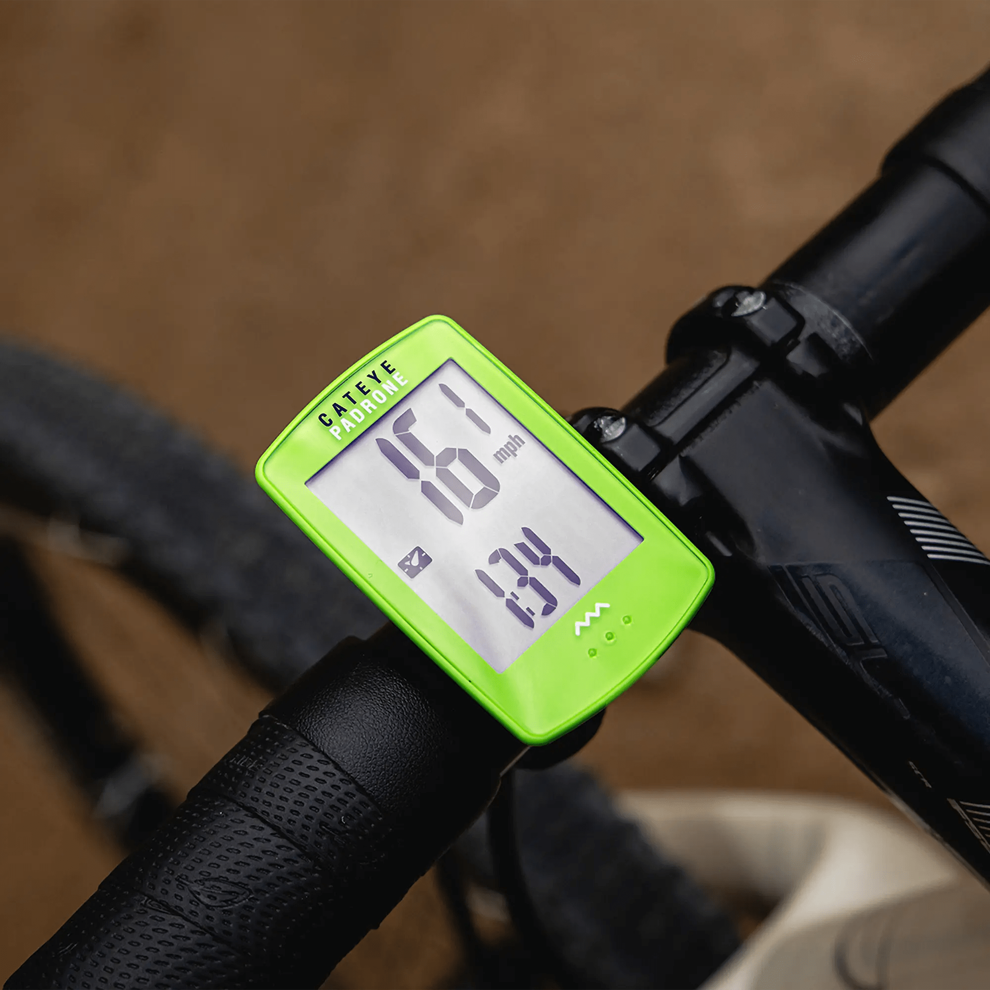 A close-up of a green CATEYE Padrone (CPSC) bike computer with a large display, mounted on a handlebar, shows 18.1 mph and 2.34 miles, with part of the bicycle and ground visible in the background.