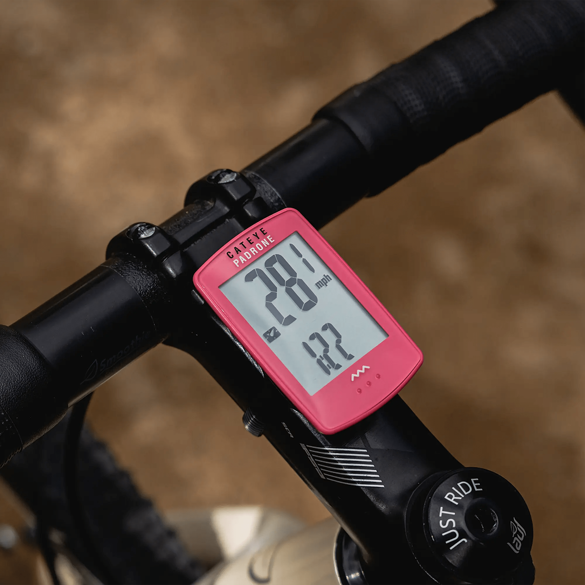 A red CATEYE Padrone (CPSC) bike computer with a large display is mounted on a handlebar, showing 28.1 mph speed and 1.2 miles distance against a blurred brown terrain background.