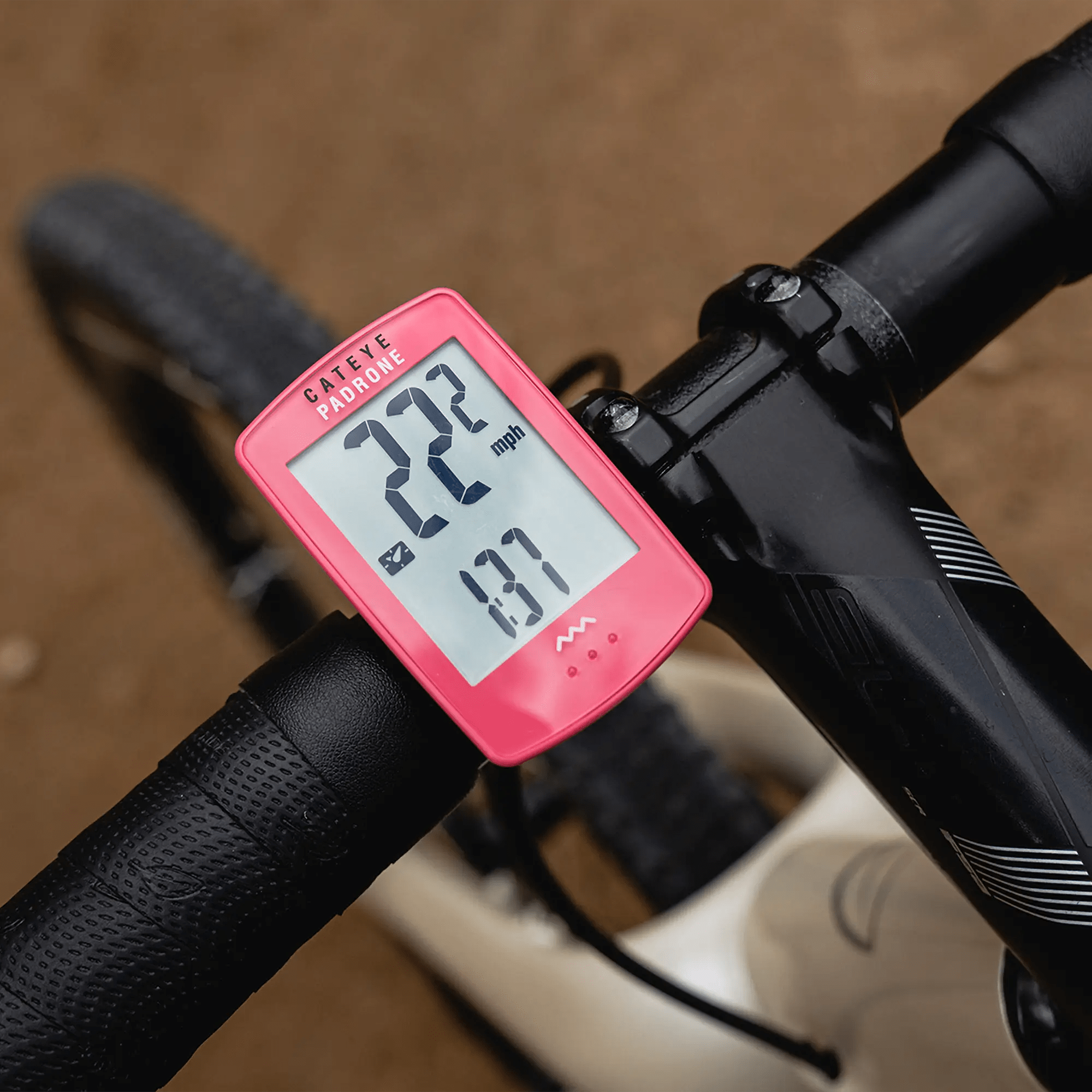 A close-up of a pink CATEYE Padrone (CPSC) bike computer mounted on black handlebars displays 22.2 mph and 1.37 distance, with a blurred dirt path in the background.