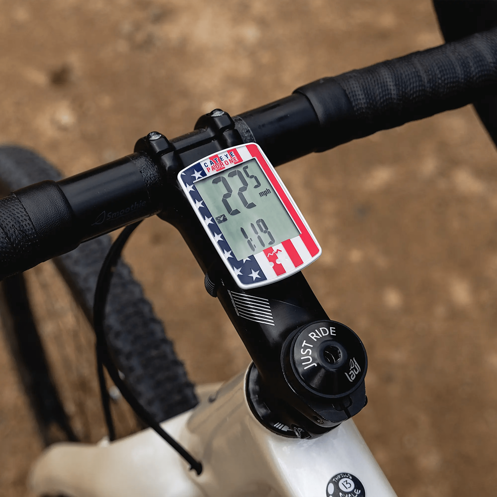 A CATEYE Padrone (CPSC) bike computer with an American flag design and large display reads 22.3 mph and 1.19 miles, mounted on a black handlebar of a white bike on a dirt path.