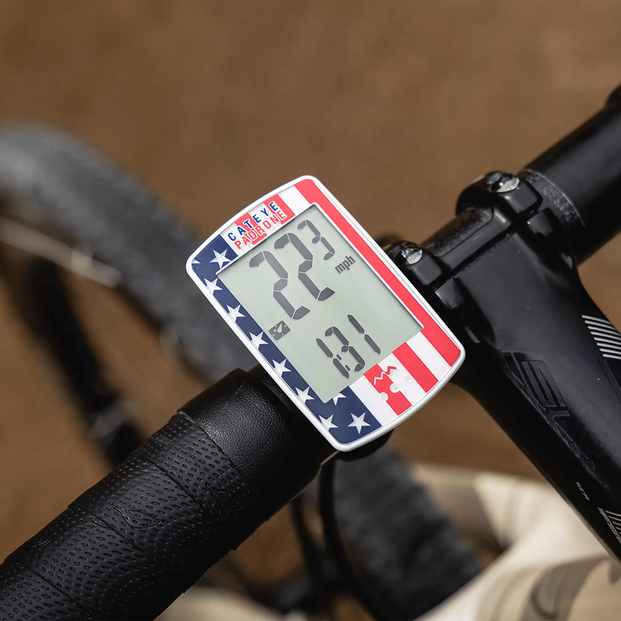 A CATEYE Padrone (CPSC) bike speedometer with an American flag design and large display shows 22.3 mph and 0.1, mounted on the handlebars of a black bicycle.