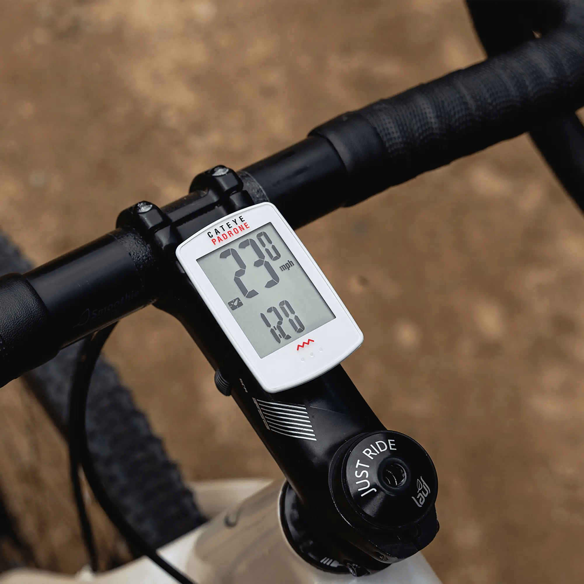 A close-up of a bike handlebar with the CATEYE Padrone (CPSC) and its large display showing 23.9 mph speed and 1.20 miles distance, controlled by the ClickTec™ interface, with a dirt path in the background.