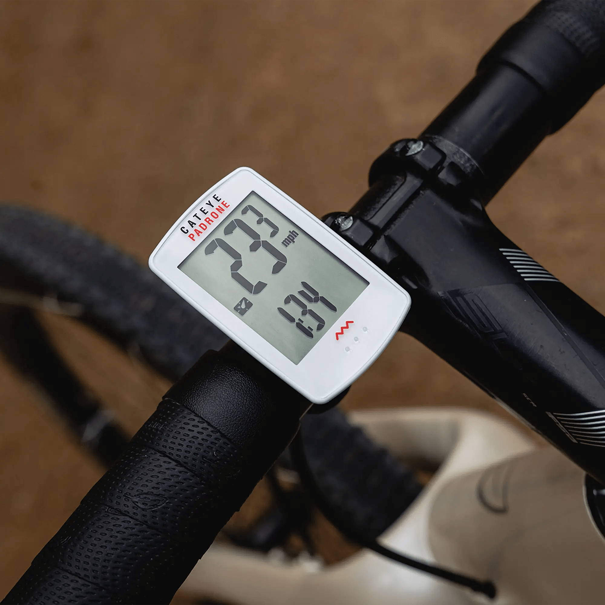A close-up of a CATEYE Padrone (CPSC) bike speedometer on a handlebar shows 23.3 km/h speed and 1.34 km trip distance. The large display stands out as the black and white bike moves over blurred brown terrain.