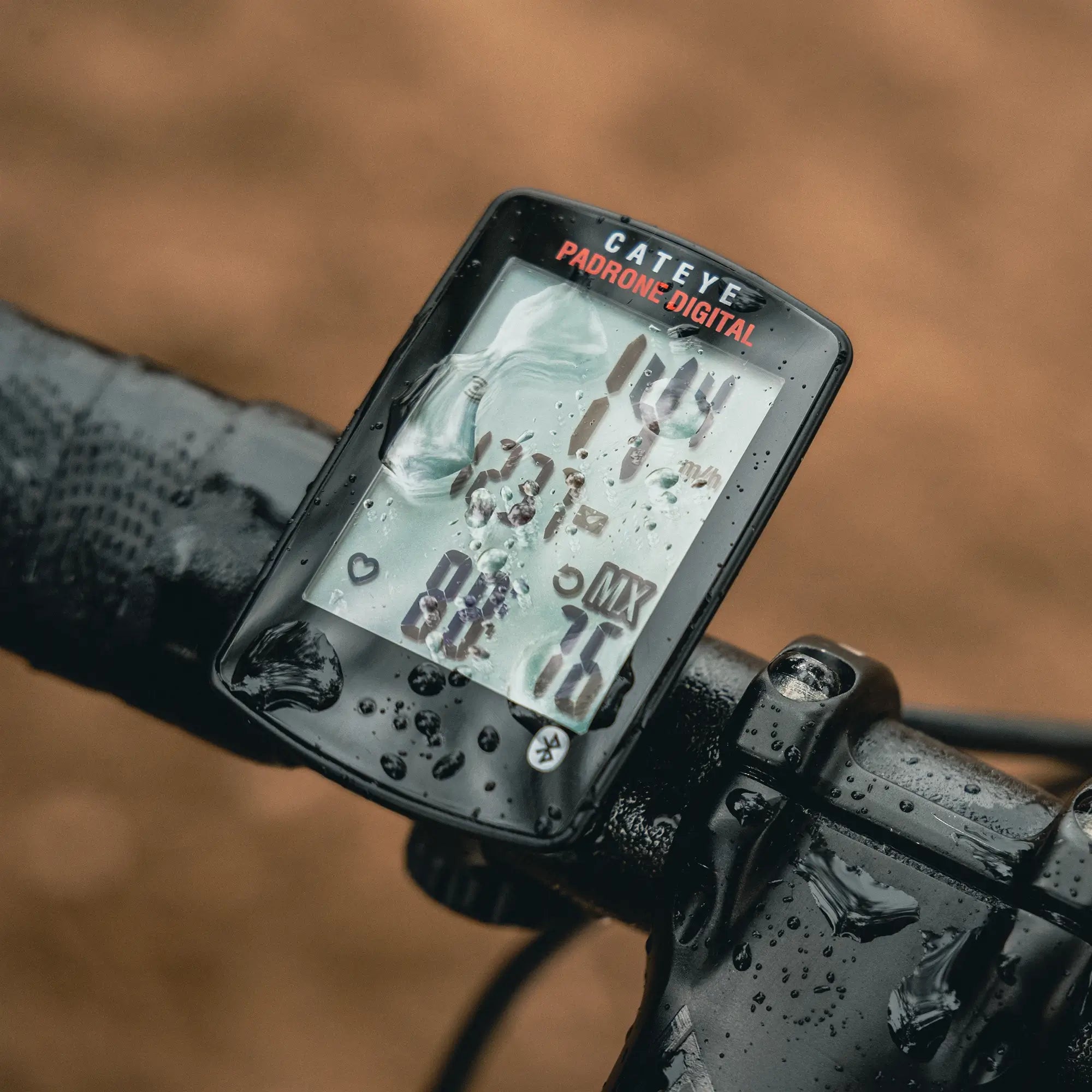 Close-up of a wet CATEYE Padrone Digital (CPSC) bike computer with wireless sensors, mounted on handlebars and showing cycling data, raindrops visible on the screen, and a blurred brown background.