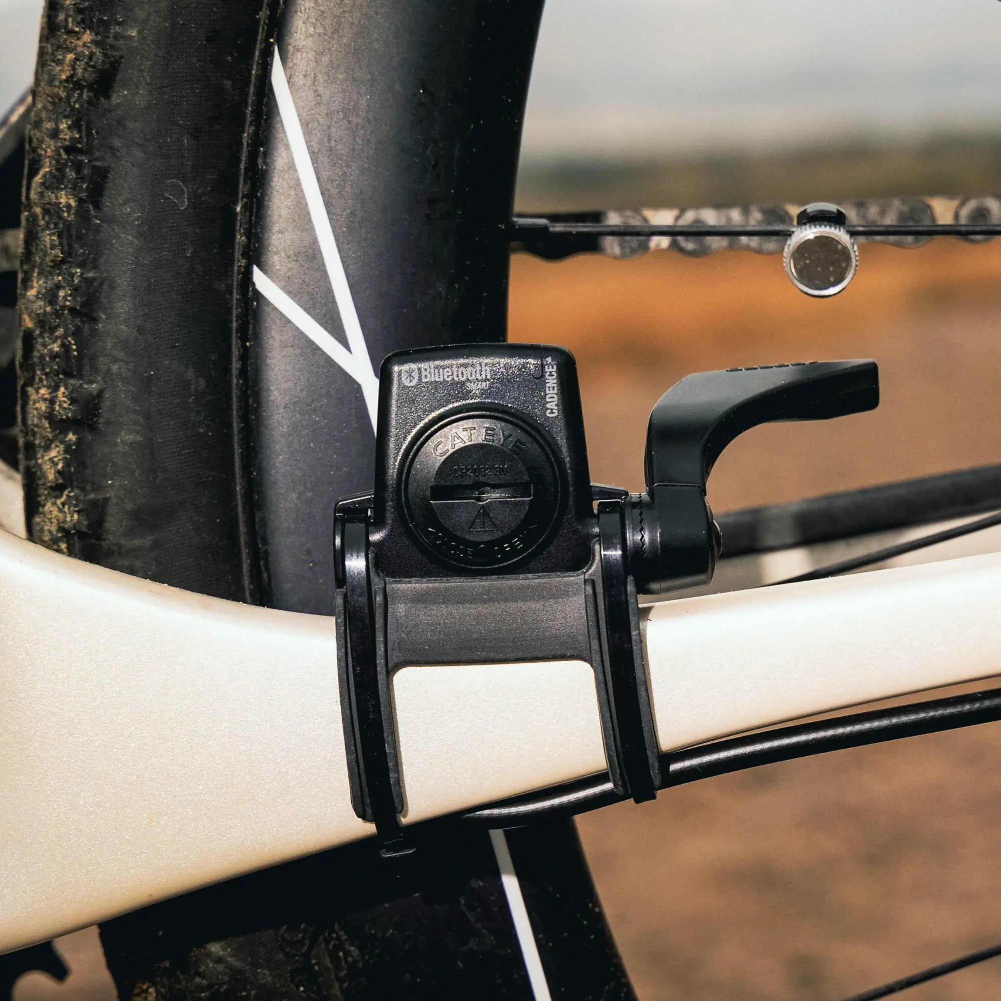 Close-up of a CATEYE Padrone Digital (CPSC) black Bluetooth cadence sensor attached to a bike frame, delivering accurate cycling data with wireless sensors. Part of the rear wheel, tire, and chain appears in the background.