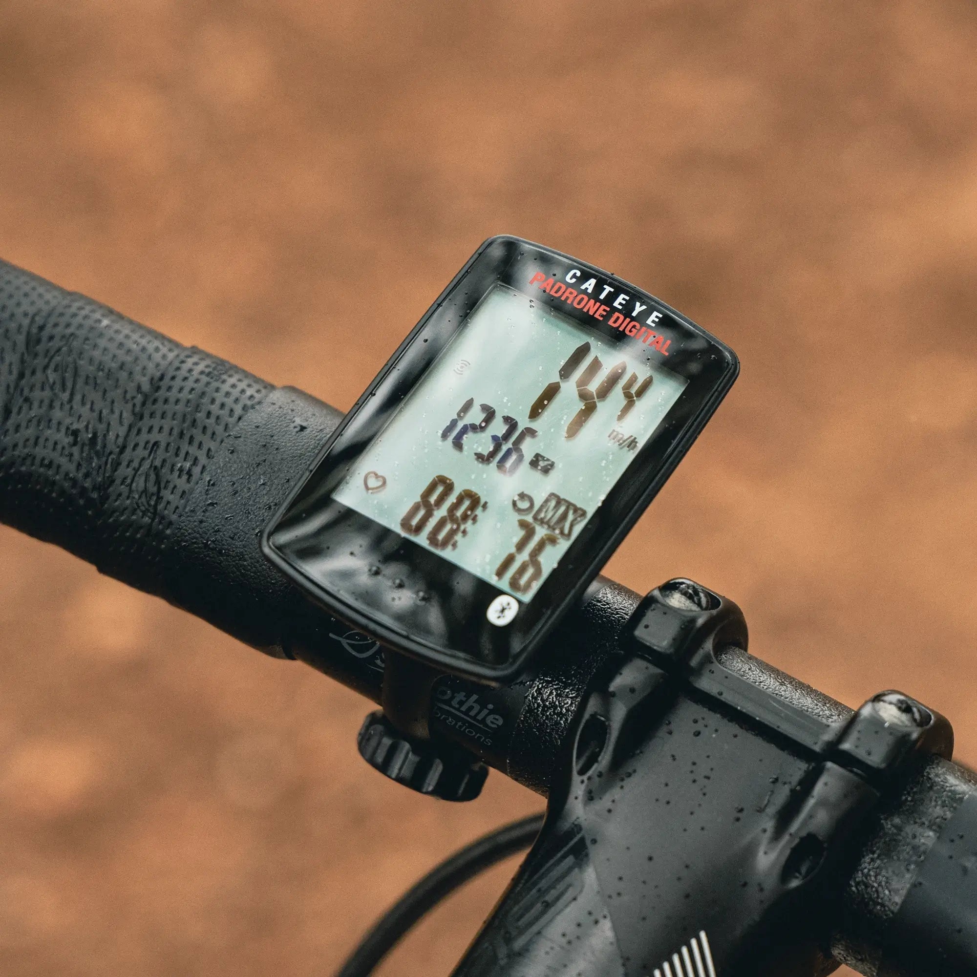 A close-up of a CATEYE Padrone Digital (CPSC) bike computer with wireless sensors on handlebars, showing speed (14.4 km/h), time (12:36 PM), and other cycling data against a blurred brown background.