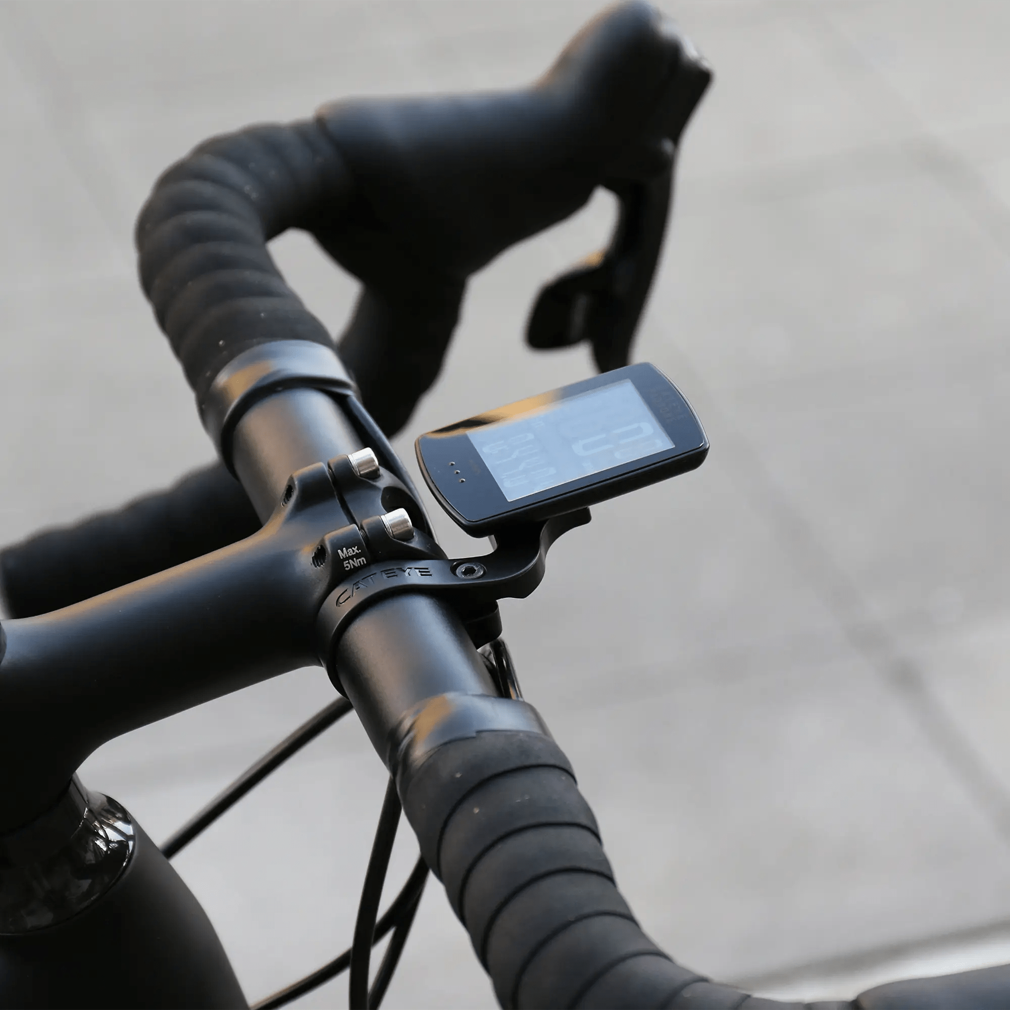 Close-up of a bicycle handlebar with wrapped grips, featuring the CATEYE Padrone Stealth (CPSC) cycling computer with an inverted LCD, securely mounted on an OF-100 Out-front bracket against a blurred light gray pavement background.