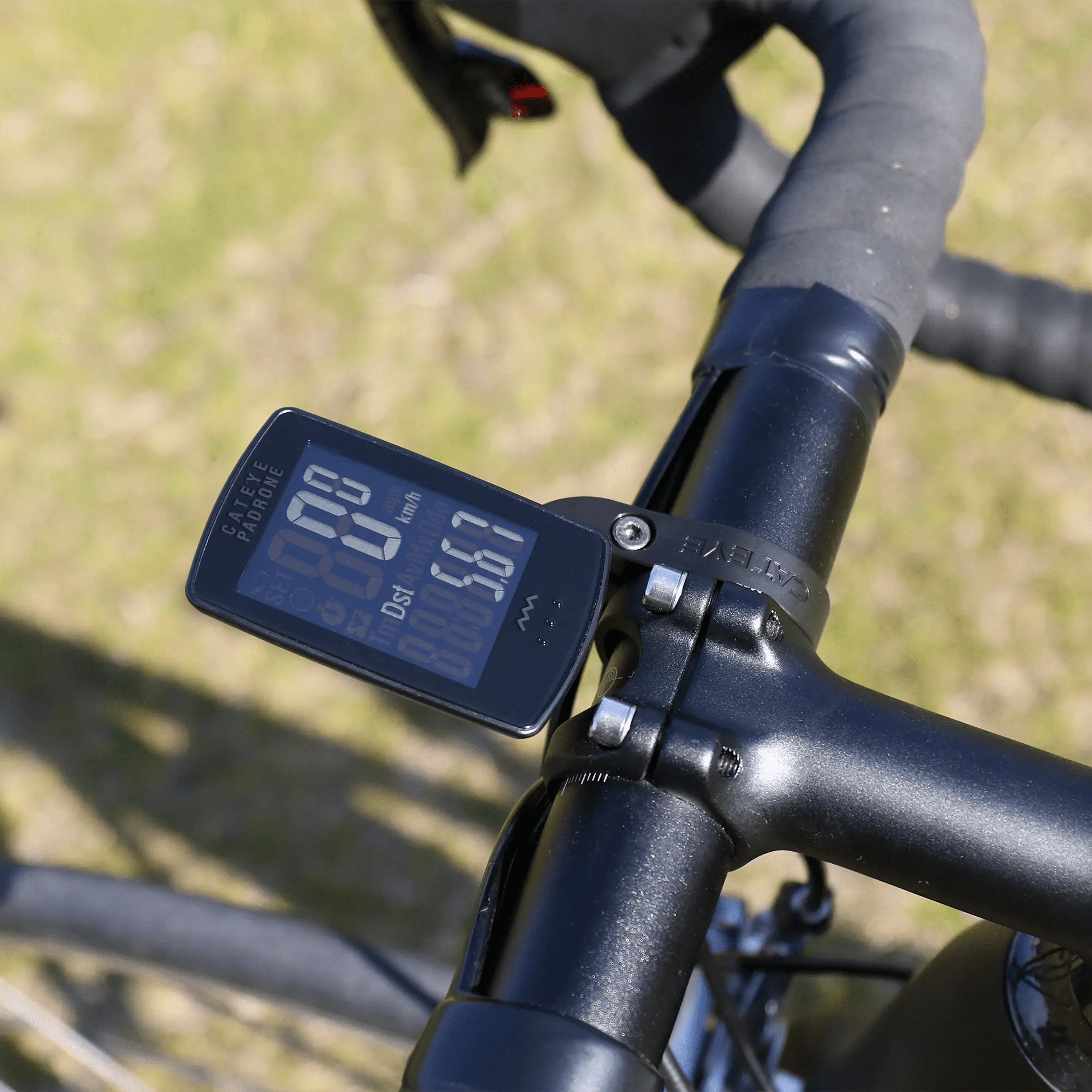 A close-up of a bicycle handlebar with the CATEYE Padrone Stealth (CPSC) bike computer, featuring an inverted LCD showing speed, distance, and ride time. It’s mounted near the stem with the OF-100 Out-front bracket against a grassy backdrop.