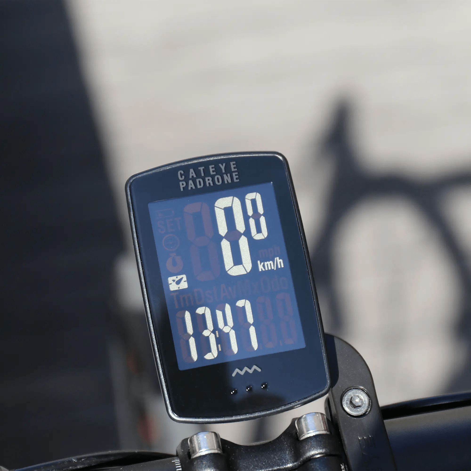 A close-up of a CATEYE Padrone Stealth (CPSC) bike computer with inverted LCD, mounted on handlebars via the OF-100 Out-front bracket, displays 0 km/h and 13:17, with a blurred background and wheel shadow visible.