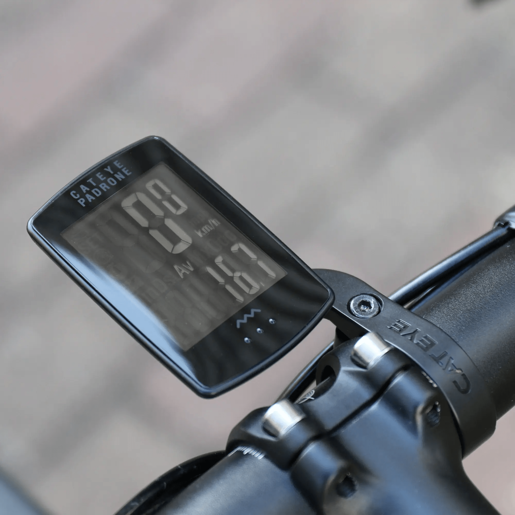 A CATEYE Padrone Stealth (CPSC) bike computer with an inverted LCD is mounted on a bicycle handlebar, showing speed and average speed in kilometers per hour.