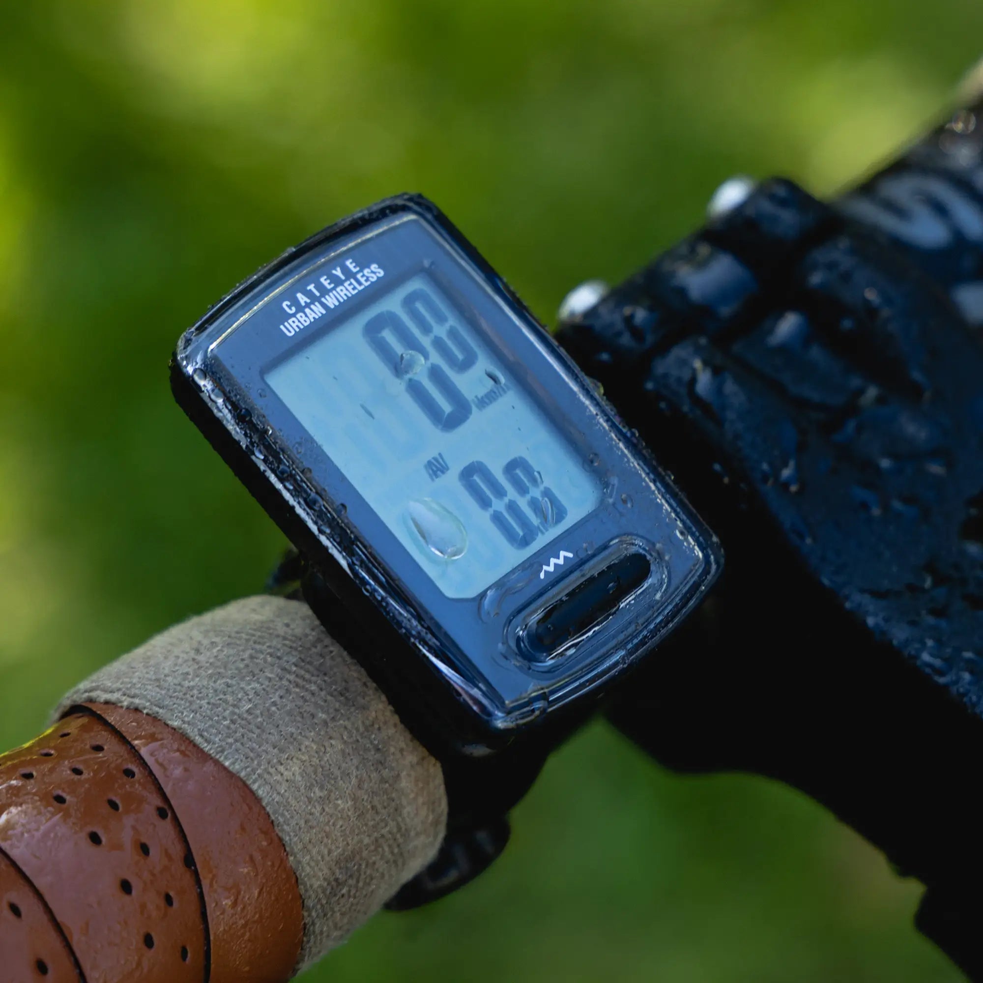 A close-up of a CATEYE Urban Wireless (CPSC) bike computer with a programmable odometer, mounted on a FlexTight™ universal bracket beside a leather-wrapped grip, set against a blurred green background.
