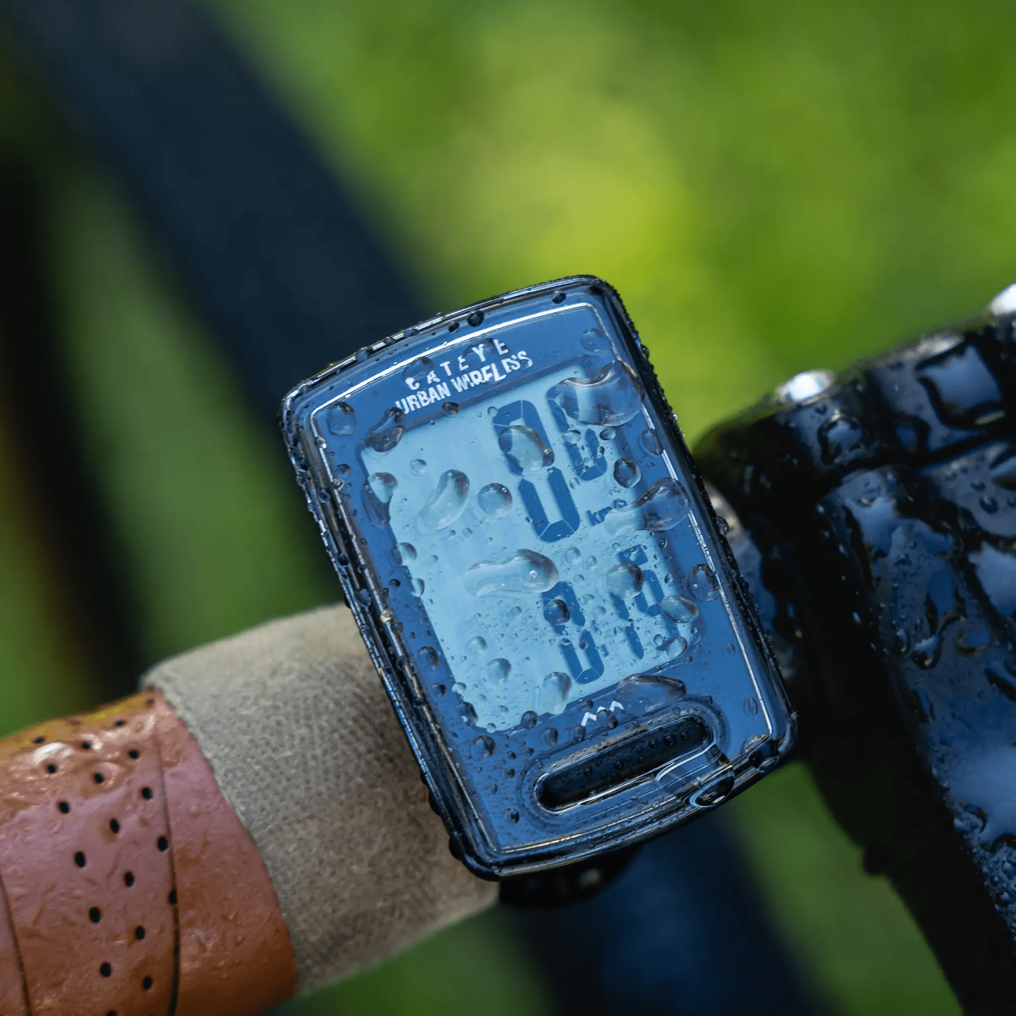 A close-up of a wet CATEYE Urban Wireless (CPSC) bike computer mounted on handlebars with a FlexTight™ bracket, showing digital speed and distance. Water droplets dot the screen, while the green background is softly blurred.