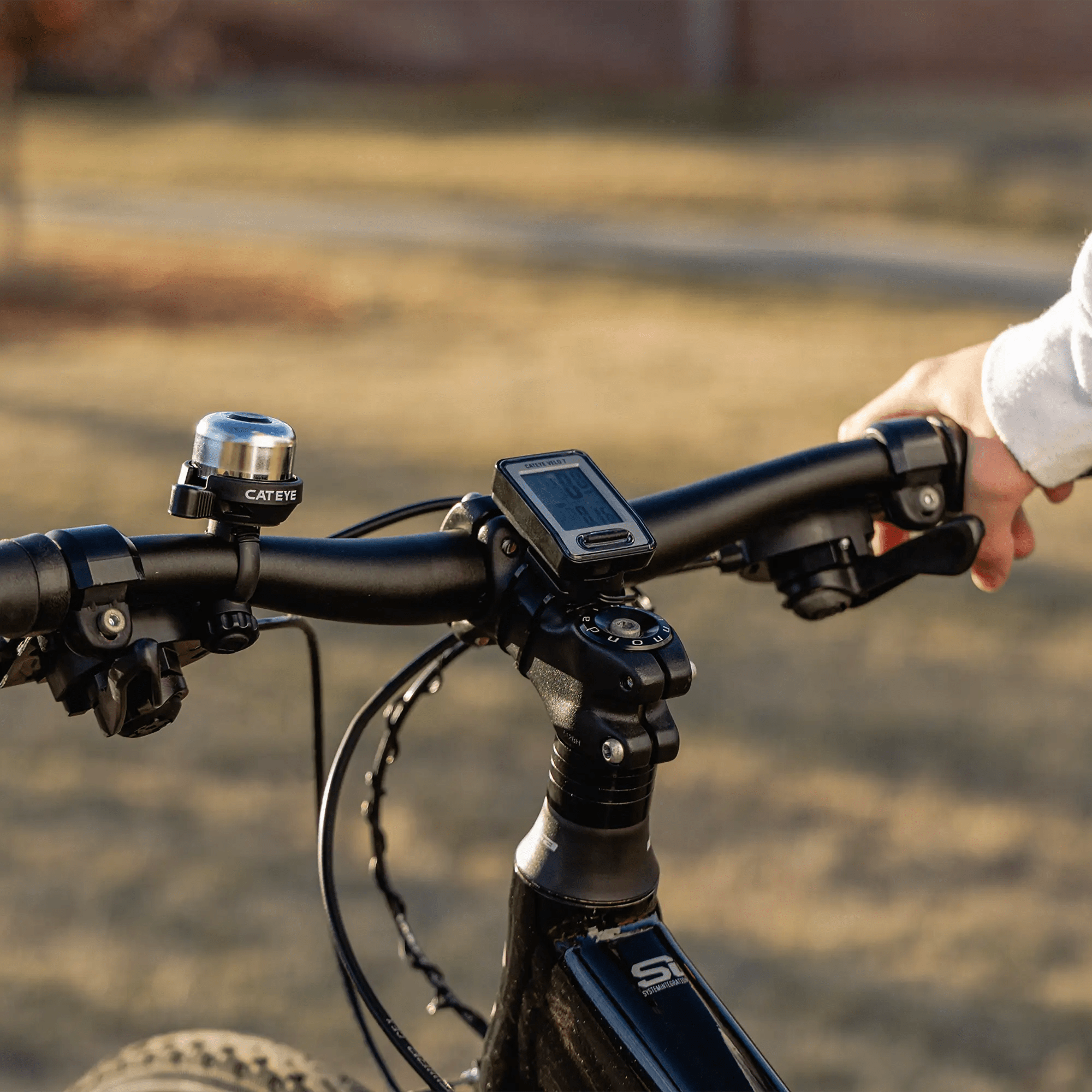 A close-up of a hand gripping an entry-level handlebar, featuring the CATEYE Velo 7 (CPSC) bike computer for precise tracking and a bell on the handlebars, set against a blurred outdoor background.