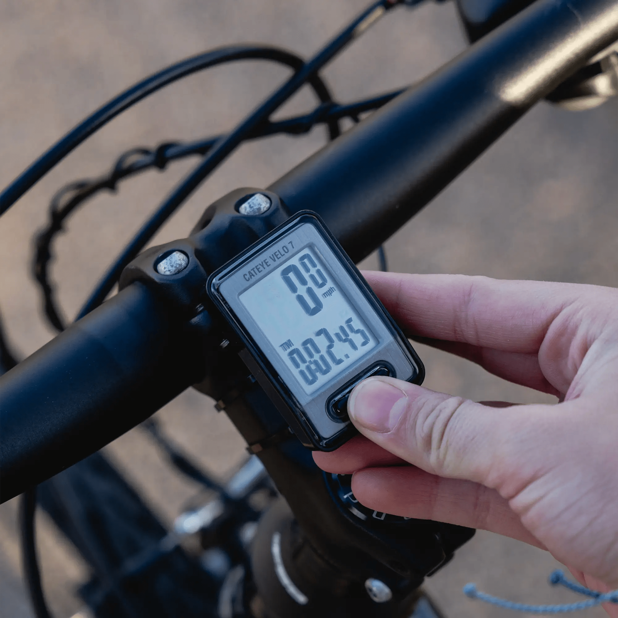 A person’s hand presses a button on the CATEYE Velo 7 (CPSC) bike computer mounted to a bicycle’s handlebars, showing precise speed and distance tracking.