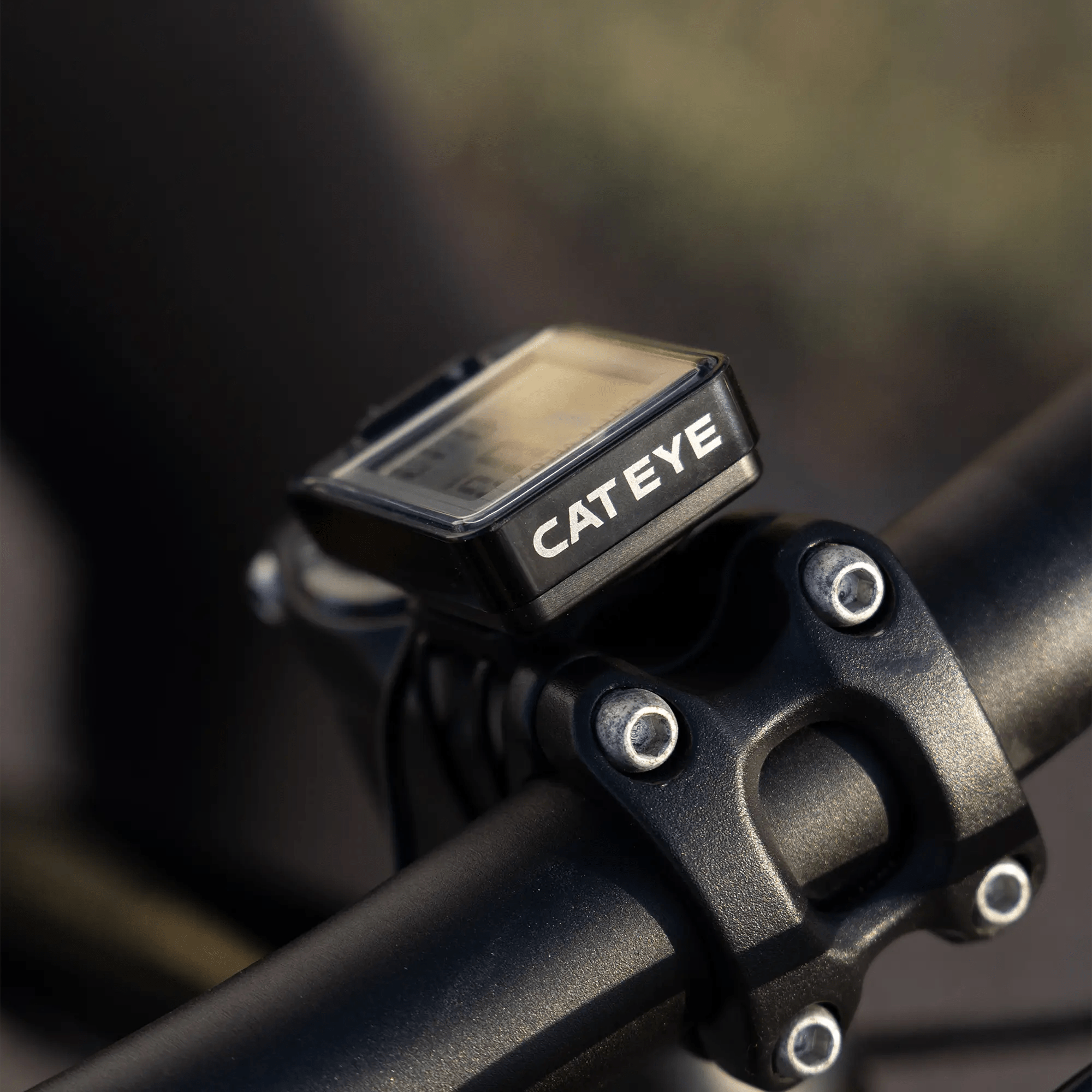A close-up of a CATEYE Velo 7 (CPSC) bike computer mounted on a bicycle handlebar, displaying its screen and the bolts securing the handlebar against a softly blurred background.
