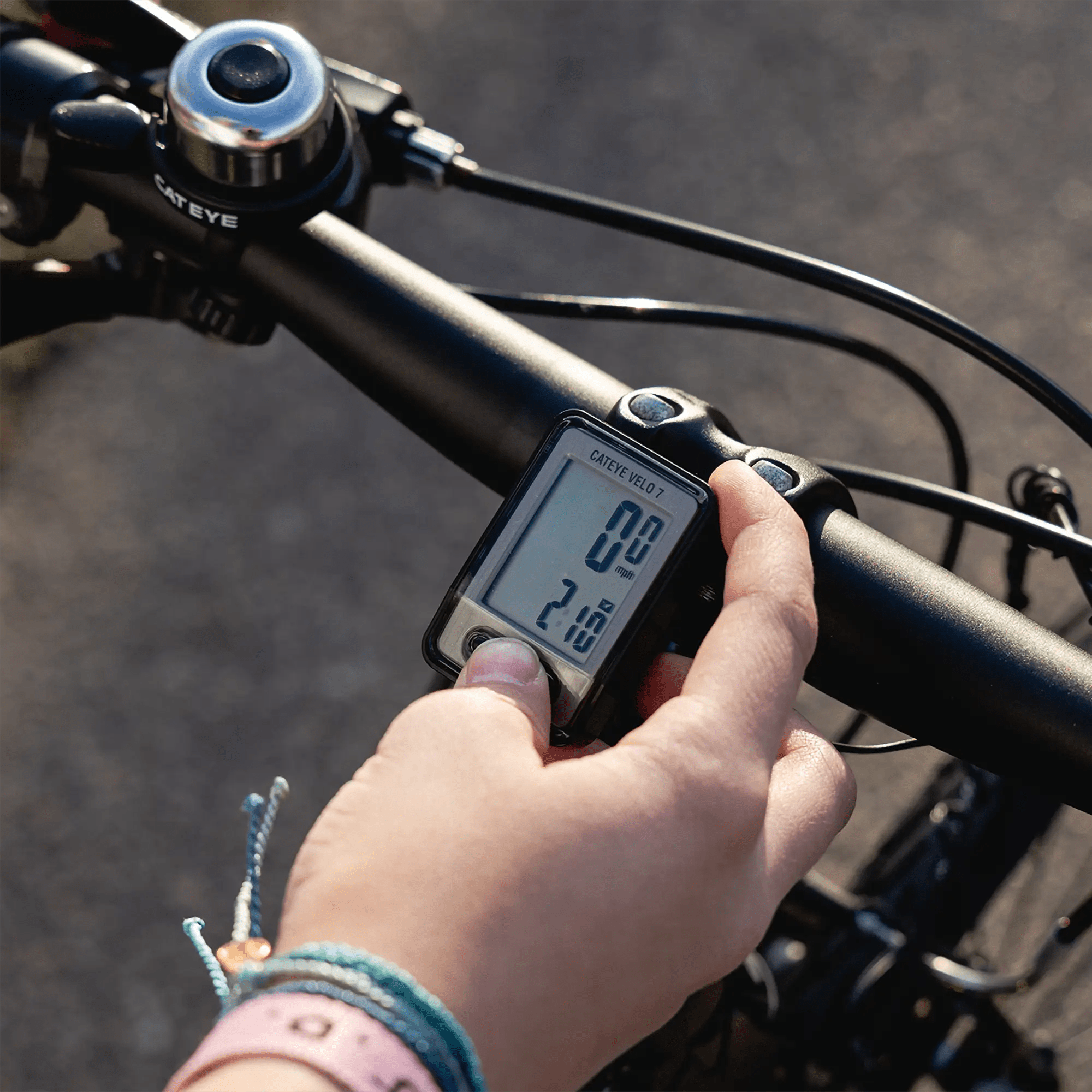 A person adjusts a CATEYE Velo 7 (CPSC) bike computer on a bicycle handlebar, which shows accurate speed and distance readings. Bracelets and a smartwatch are worn, with the outdoor pavement softly blurred in the background.