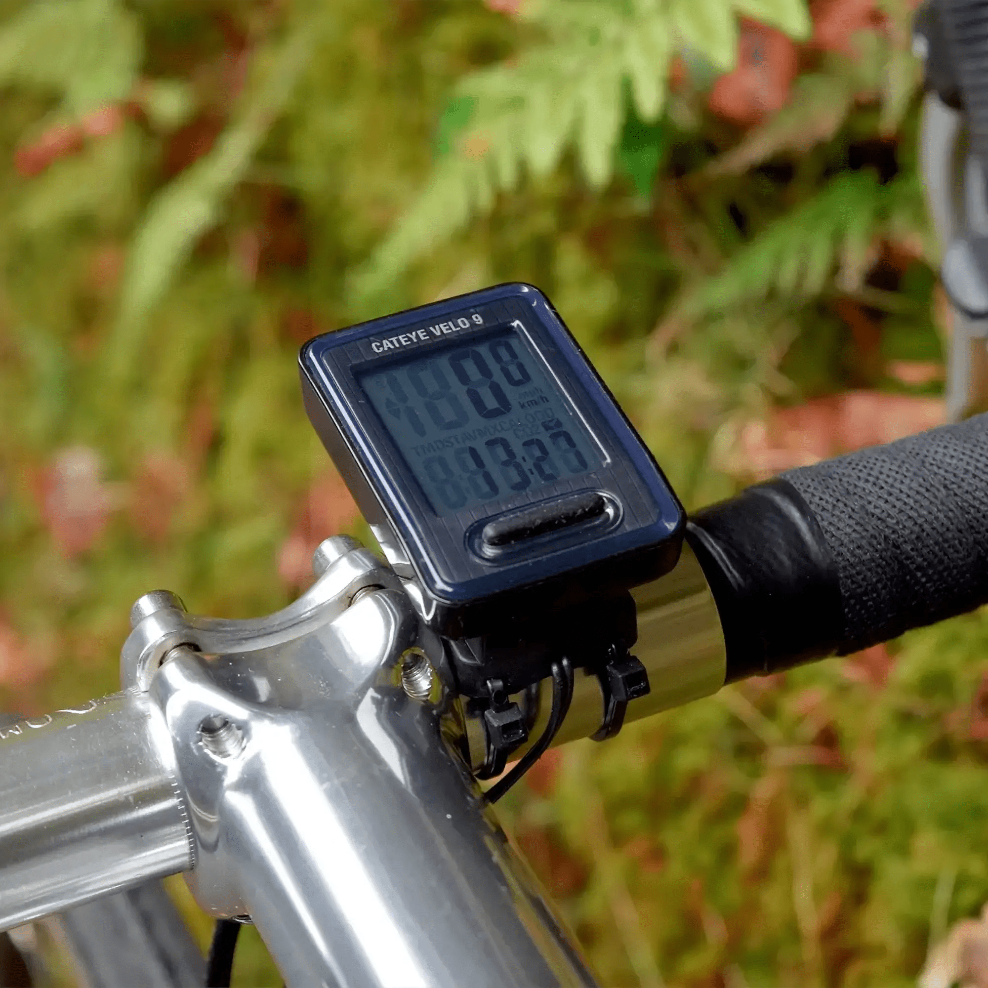 A close-up of a CATEYE Velo 9 (CPSC) bike computer mounted on handlebars, showing speed and distance against blurred green foliage—a great entry-level choice for beginner cyclists.