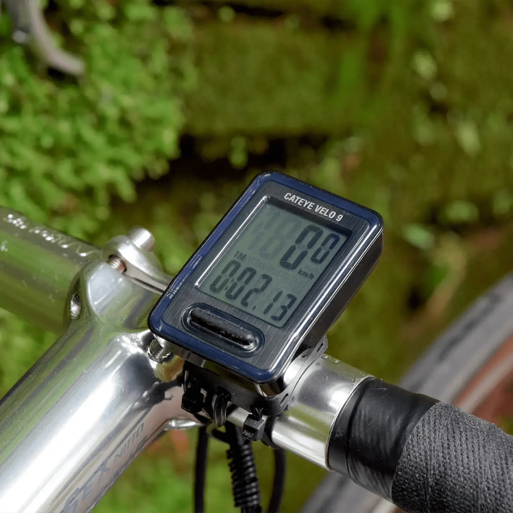 A close-up of a bicycle handlebar features a CATEYE Velo 9 bike computer showing speed, distance, and time—perfect for beginner cyclists. The blurred background is filled with green foliage.