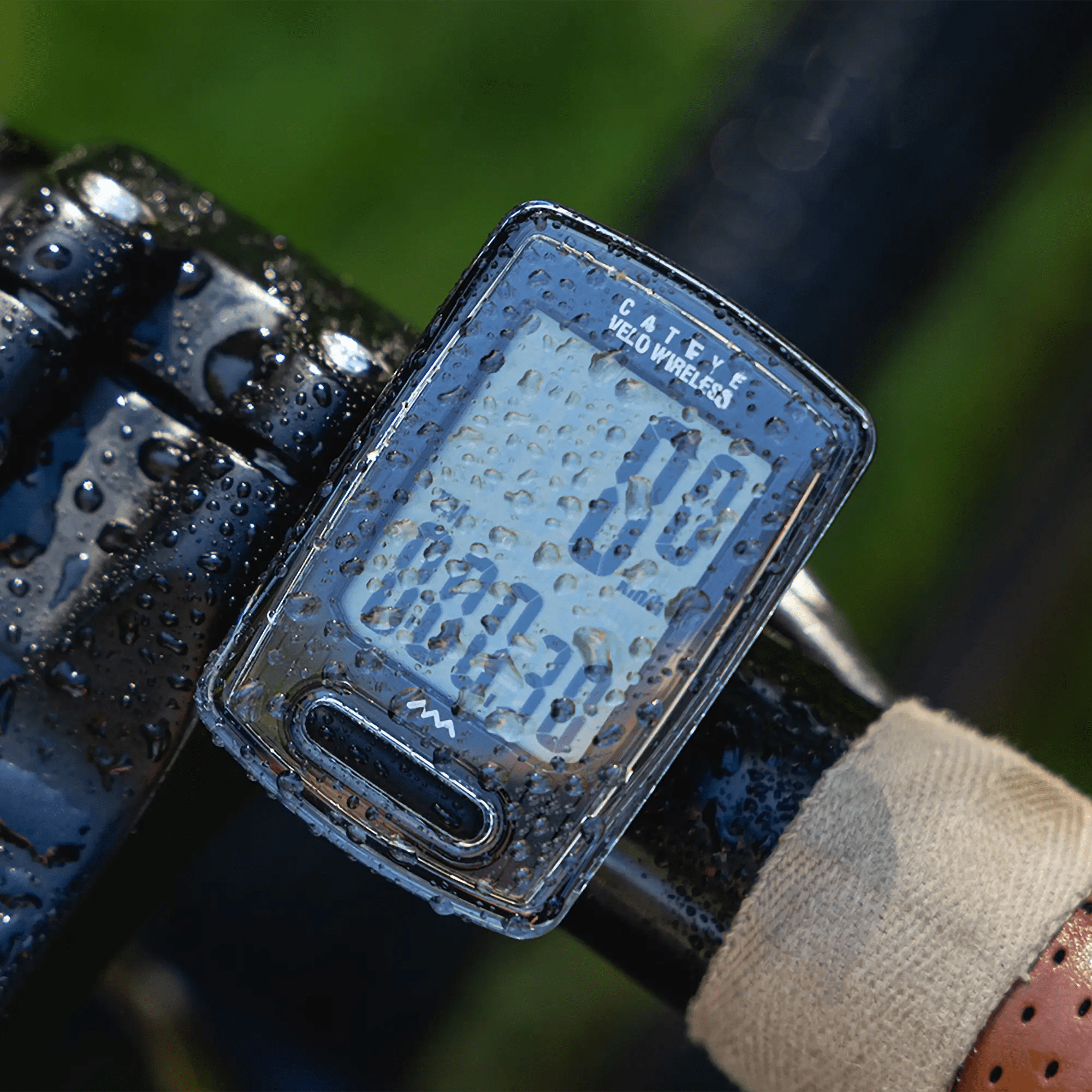 A close-up of a CATEYE Velo Wireless bike computer with programmable odometer, covered in water droplets, displays speed and distance on a beige handlebar against a green blurred background.