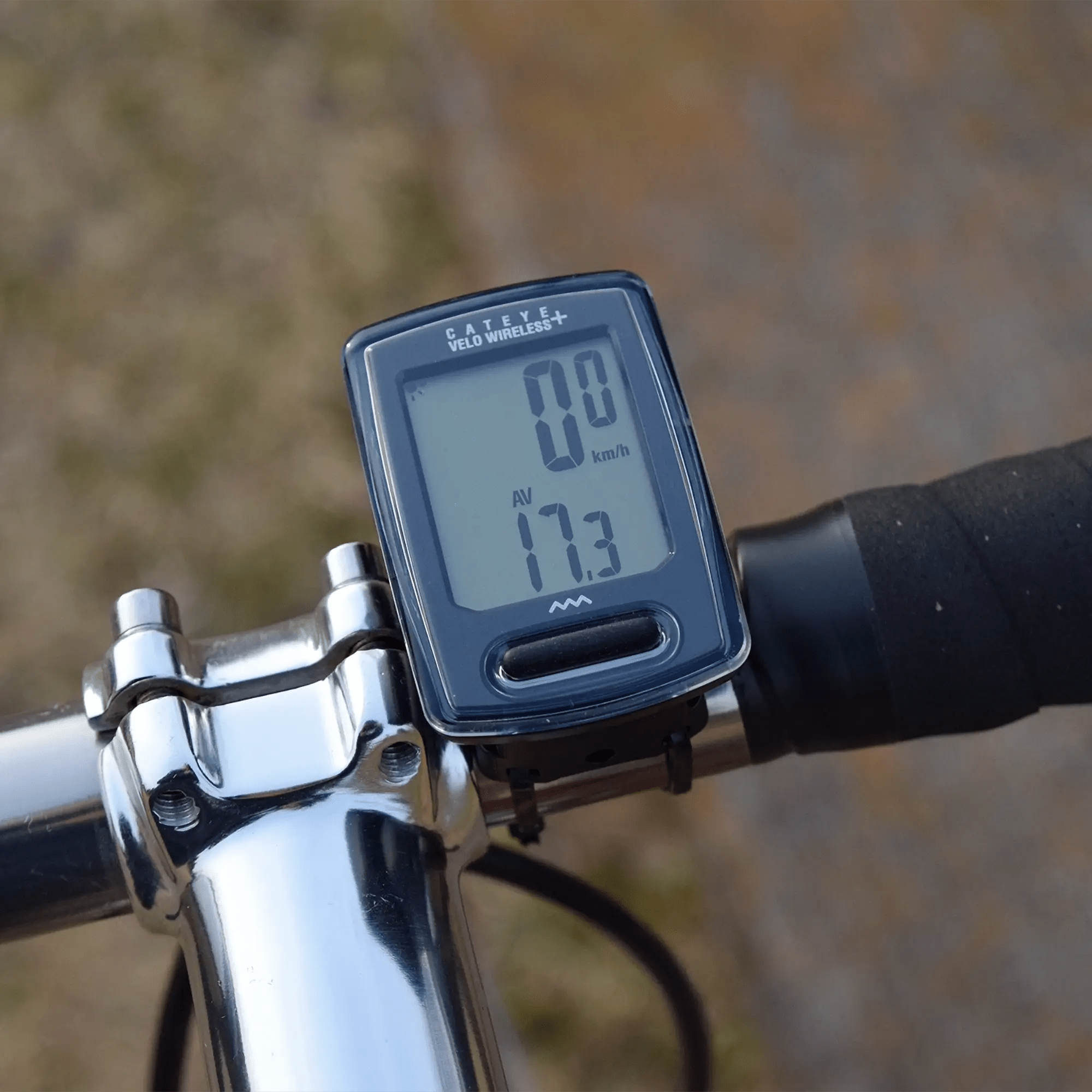 Close-up of a CATEYE Velo Wireless+ (CPSC) bike computer on a handlebar, showing 0.0 km/h current speed and 17.3 average speed on its programmable odometer. The background is blurred.
