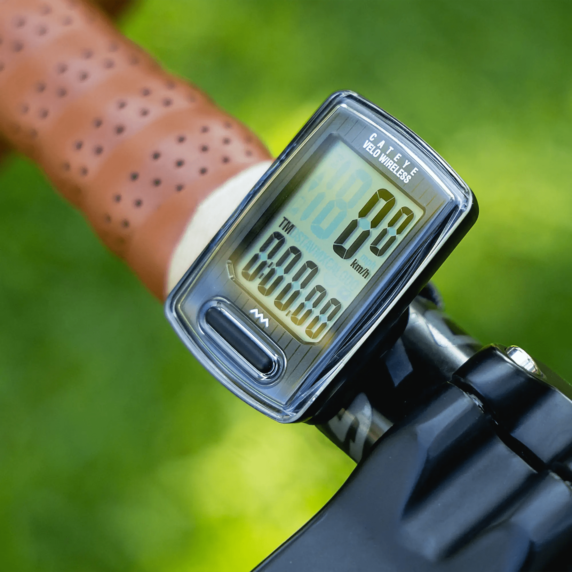 A close-up of a CATEYE Velo Wireless bicycle speedometer with a programmable odometer, mounted on handlebars with a brown perforated grip, displaying speed and distance against a blurred green background.