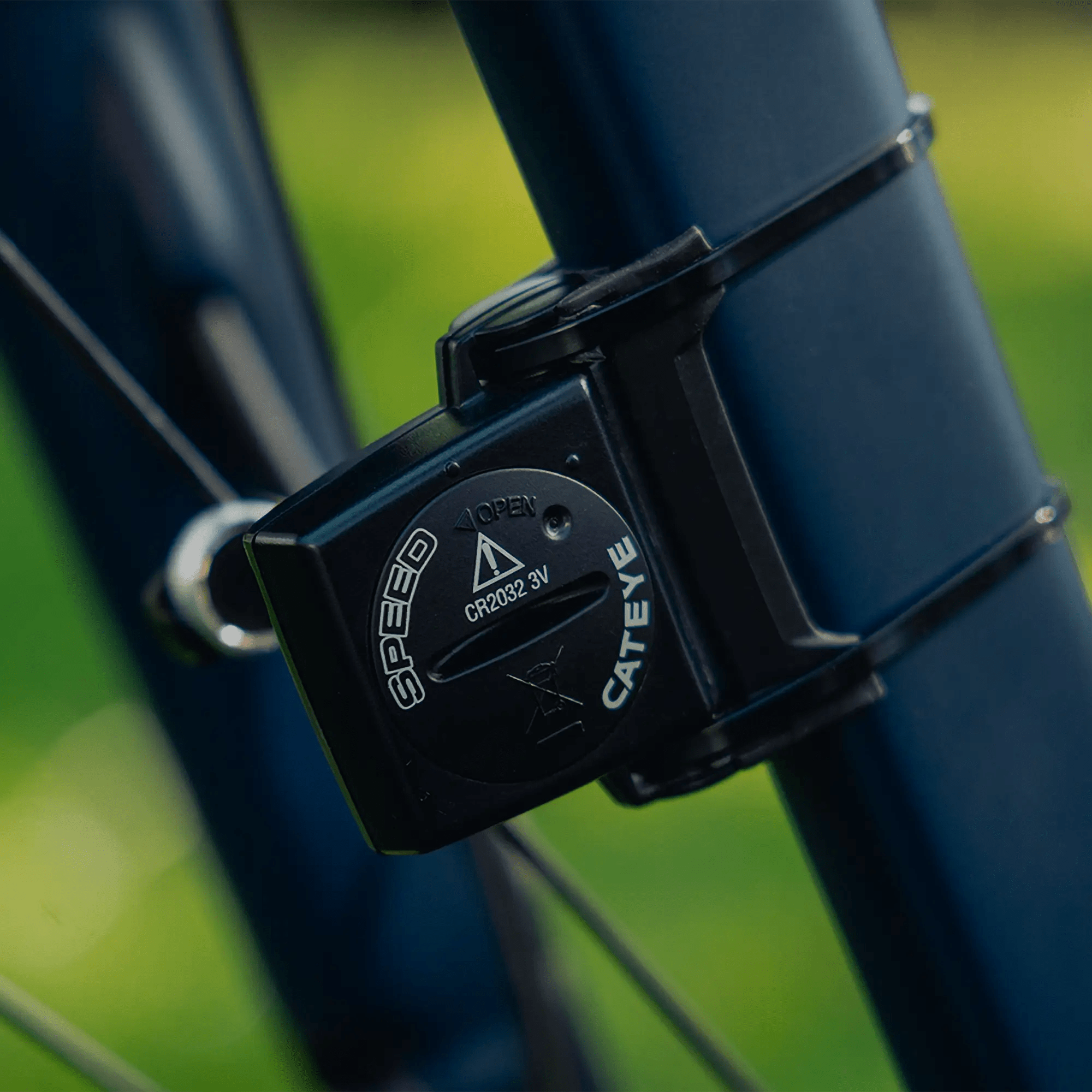 Close-up of a CATEYE Velo Wireless+ (CPSC) speed sensor attached to a bicycle frame with zip ties, featuring wireless connectivity, set against a blurred green outdoor background.