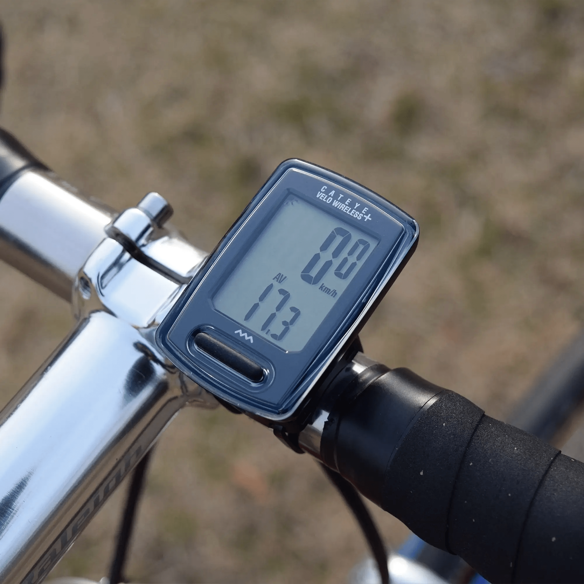 A close-up of the CATEYE Velo Wireless+ (CPSC) digital bike computer, wirelessly connected and mounted on a bicycle handlebar, displays speed and average speed against a blurred outdoor background.