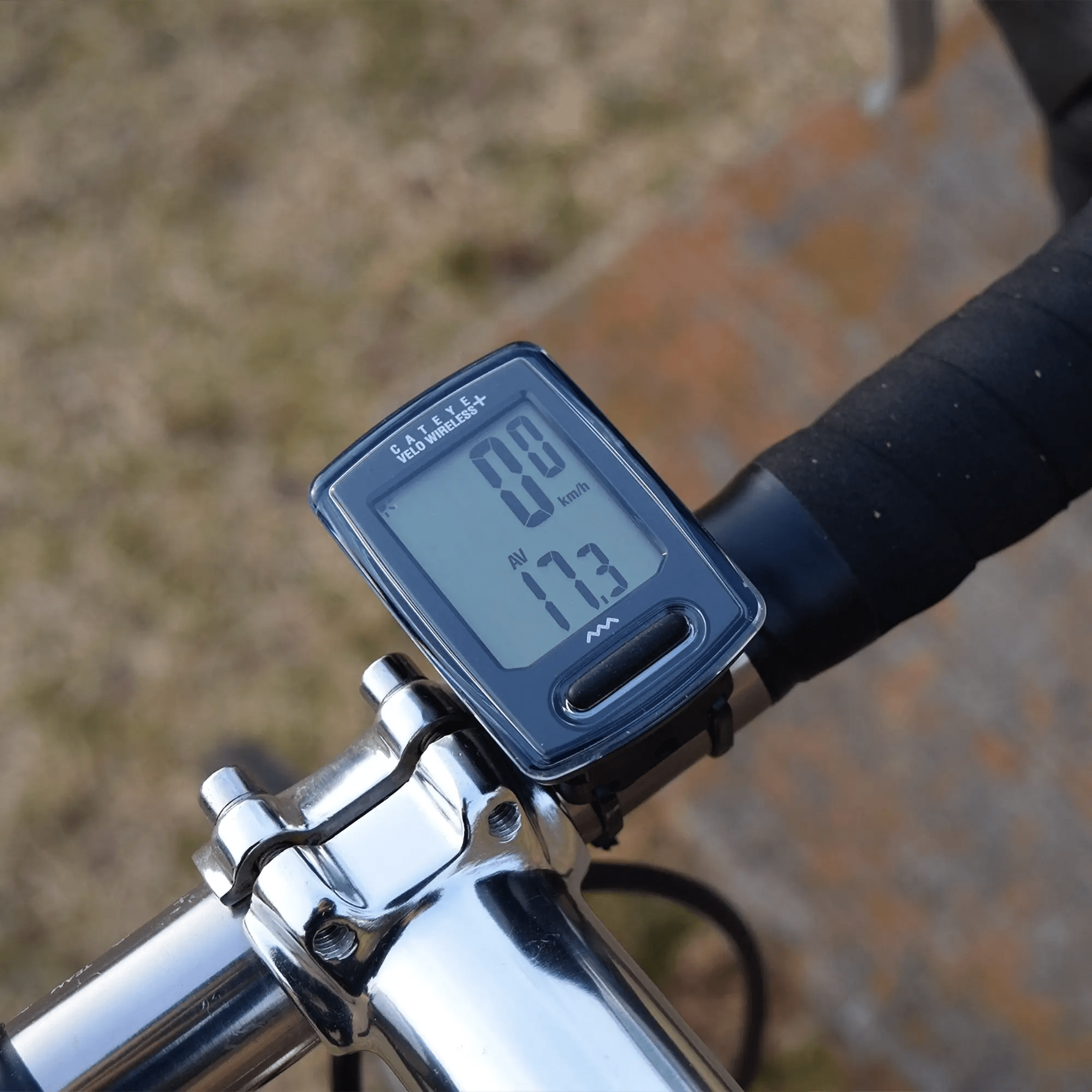A close-up of a CATEYE Velo Wireless+ (CPSC) bike computer with backlight night mode on a bicycle handlebar, showing 0.0 km/h current speed and 17.3 km/h average speed, set against a blurred outdoor background.