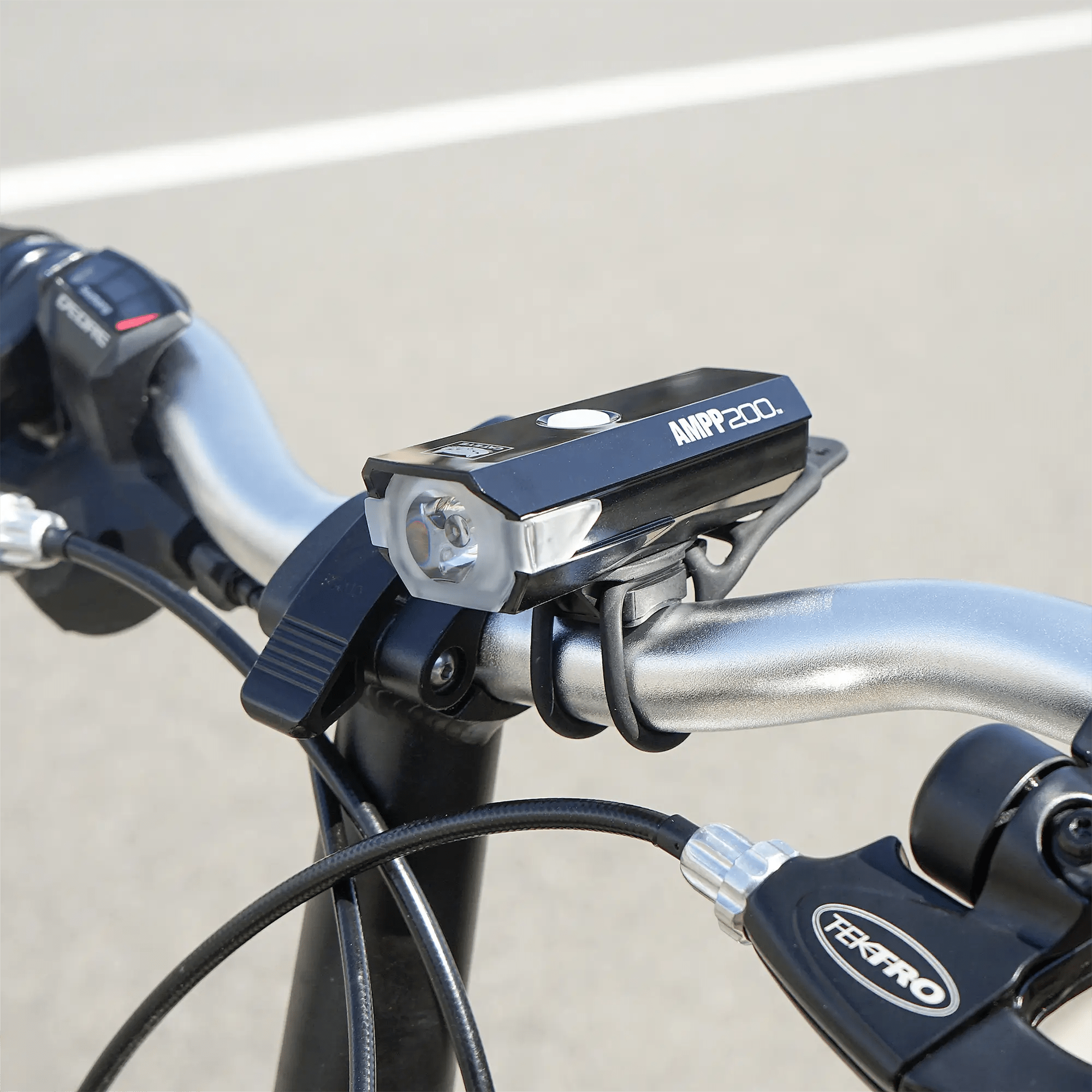 A close-up of a bicycle handlebar showcases a mounted black CATEYE AMPP200 (USB-C) rechargeable headlight with mode memory and visible brake cables, set against a blurred outdoor background.
