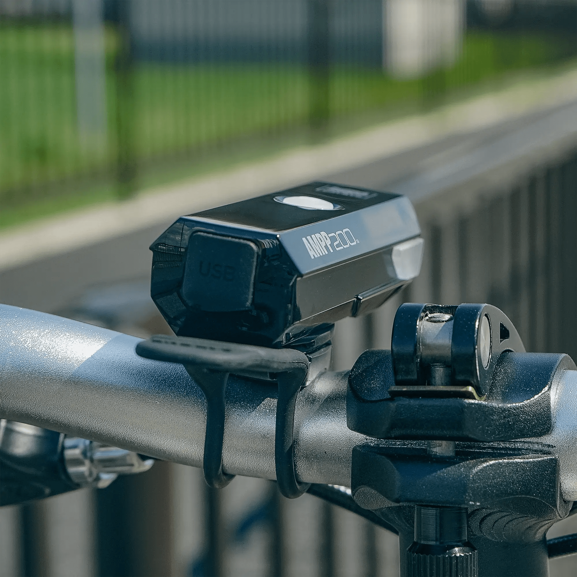 A close-up of the CATEYE AMPP200 (USB-C) bicycle headlight, delivering 200 lumens, mounted on a silver handlebar with a black rubber strap; a blurred fence and greenery appear in the background.