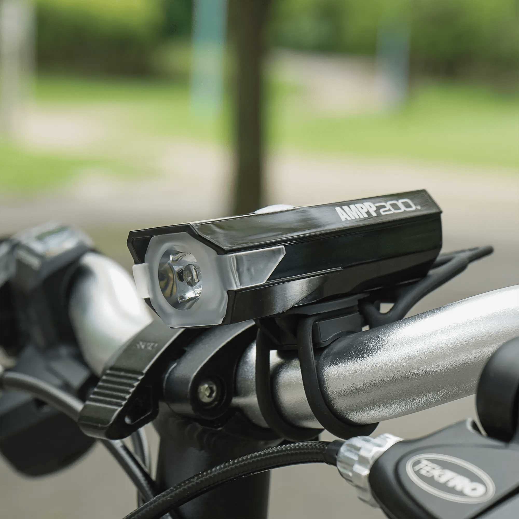 A close-up of a CATEYE AMPP200 (USB-C) 200 lumen black headlight mounted on a bicycle handlebar, set against a blurred park background.