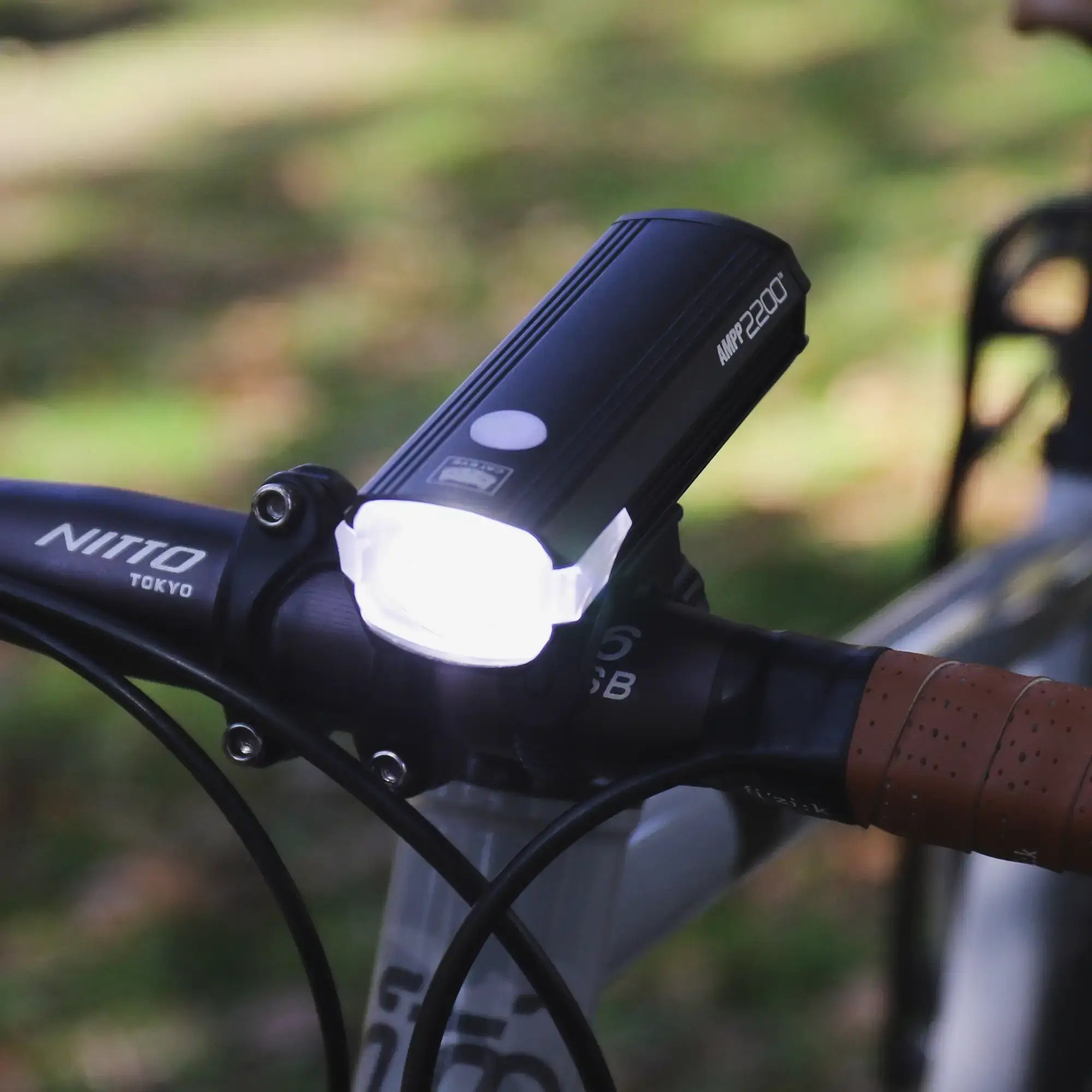A close-up of the ultra-bright CATEYE AMPP2200 bicycle headlight (2200 lumens) mounted on handlebars with leather-wrapped grips, featuring convenient USB-C charging, and shown outside in natural light.