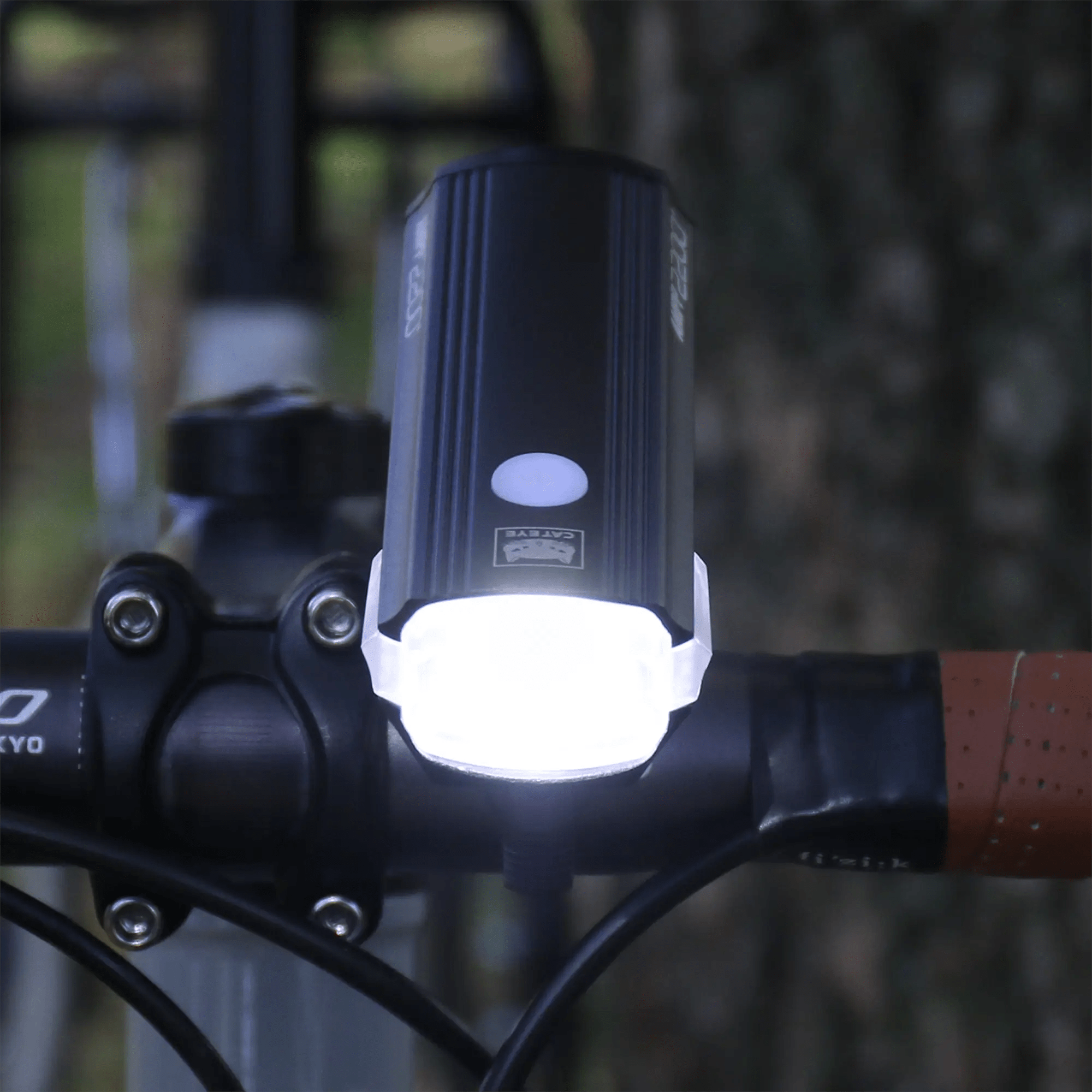 A CATEYE AMPP2200 LED bike headlight with 2200 lumens is handlebar-mounted, lighting the path ahead. A tree trunk and blurred greenery appear in the background. USB-C charging offers fast, easy recharging.