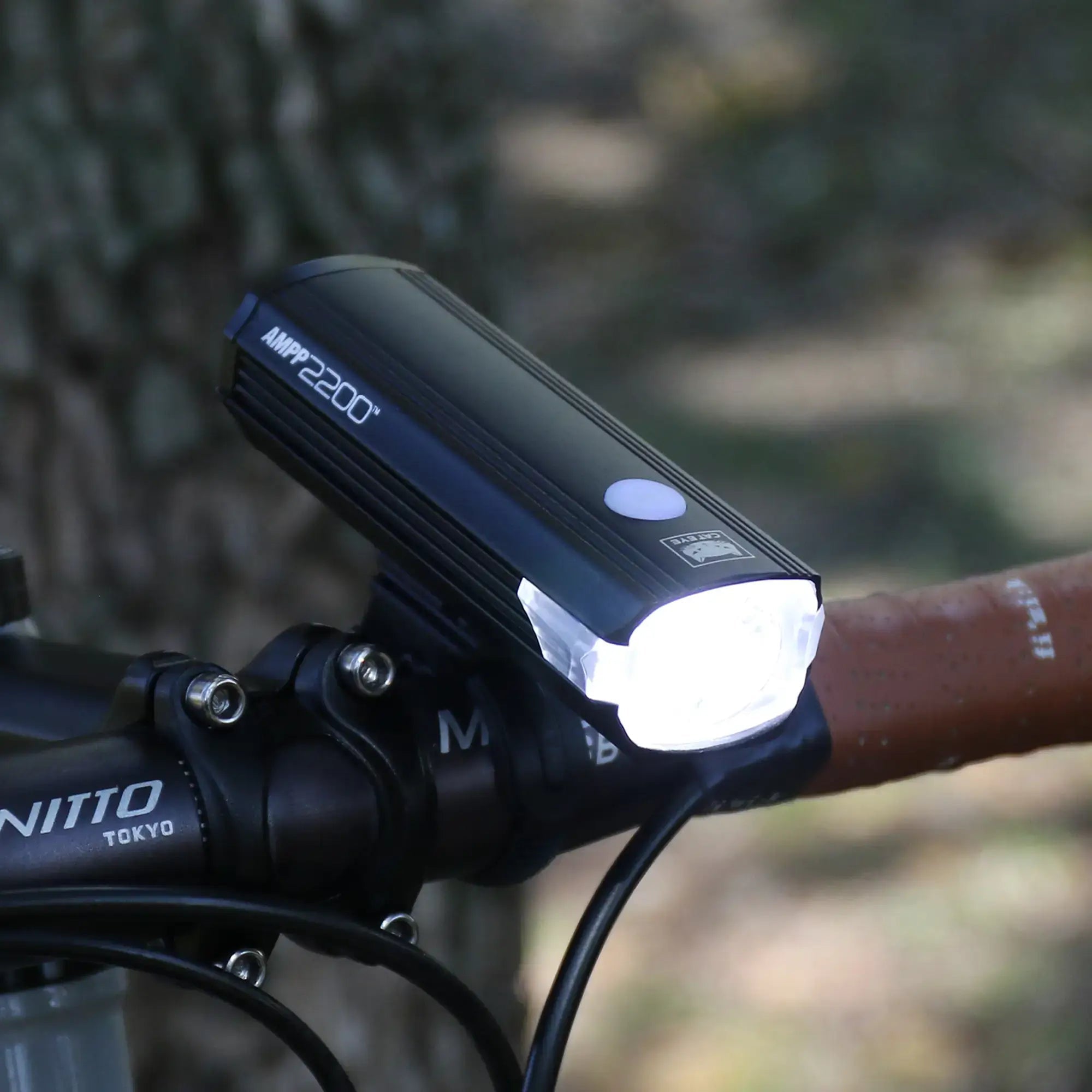 A CATEYE AMPP2200 black bicycle headlight with a 2200-lumen beam is mounted on a bike handlebar with a tan grip, shining brightly outdoors near a tree trunk.