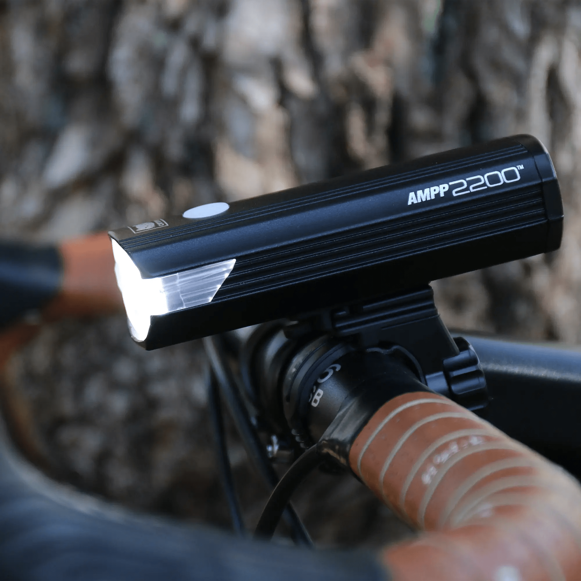 A CATEYE AMPP2200 bike headlight in black is mounted on the handlebars, emitting a powerful 2200 lumen white beam. The textured tree bark forms the backdrop, while USB-C charging ensures longer illuminated rides.