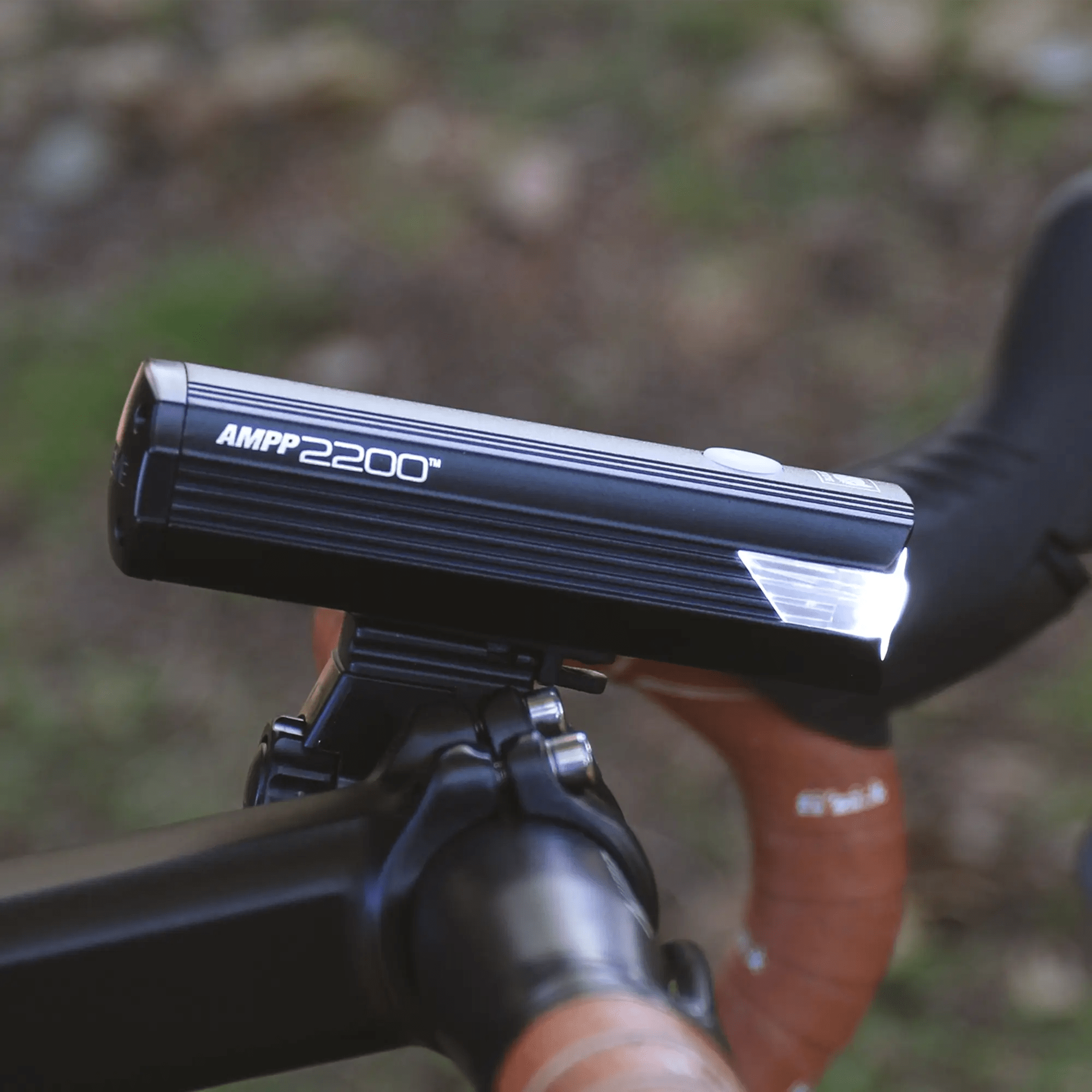 Close-up of the CATEYE AMPP2200 bike headlight mounted on handlebars, featuring a powerful 2200 lumen output and USB-C charging, shown against a blurred outdoor background.