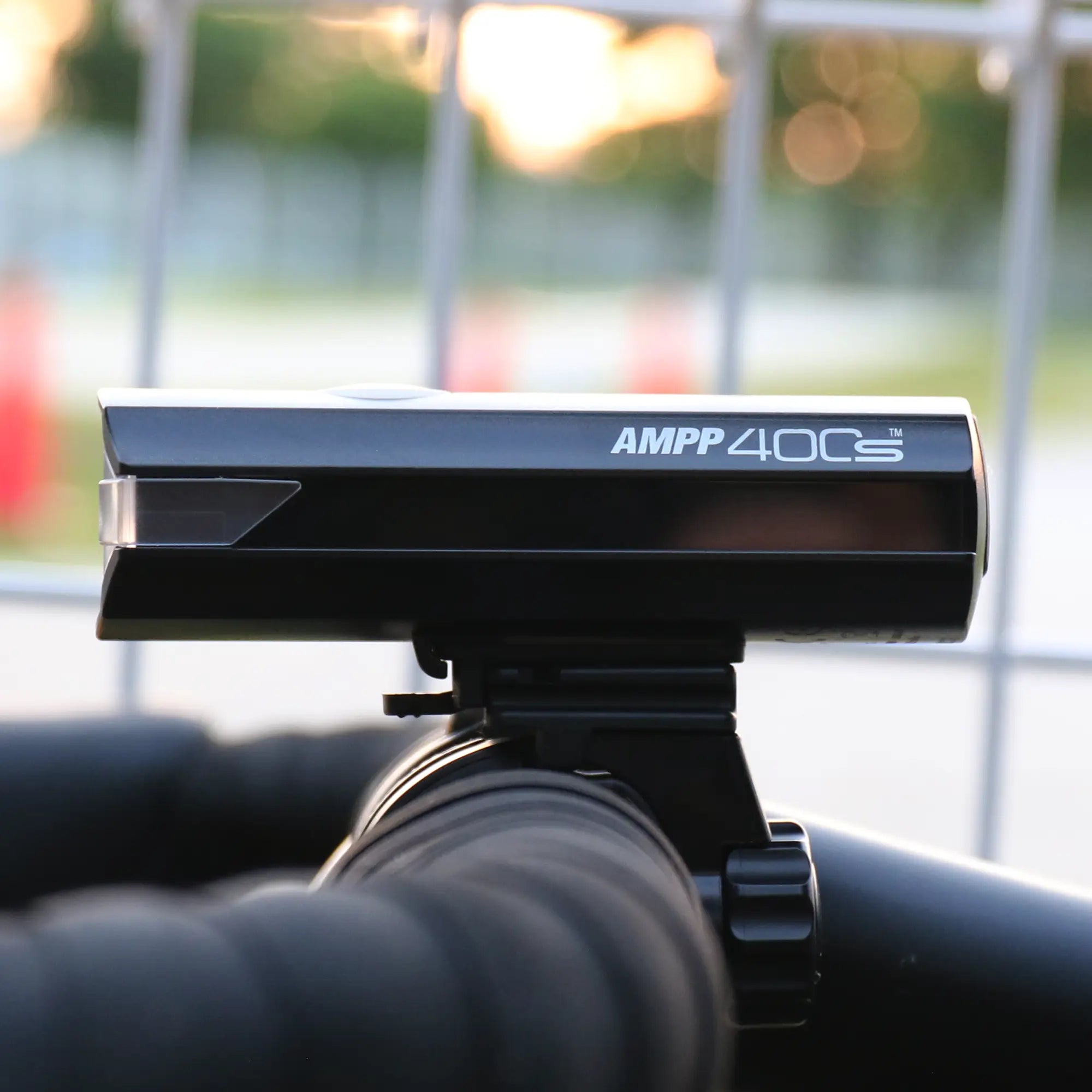 Close-up of the CATEYE AMPP400S black bike light with USB-C charging, mounted on handlebars against a blurred outdoor background and metal fence.