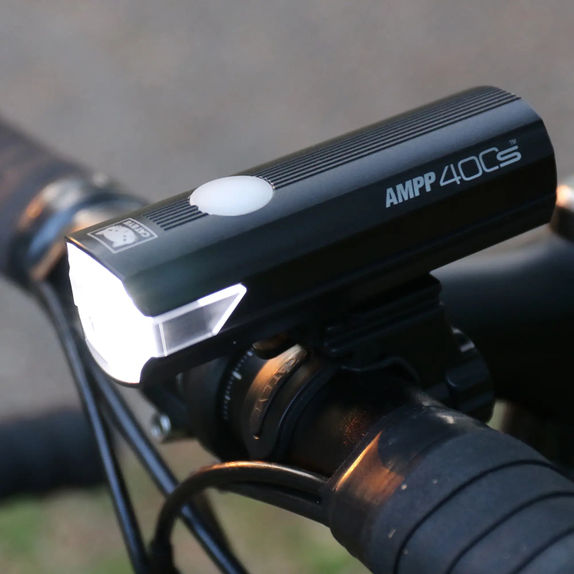 Close-up of the CATEYE AMPP400S bike light in black, mounted on handlebars and turned on, emitting a bright white beam. The USB-C charging port is visible, with handlebars and part of the bike in the background.