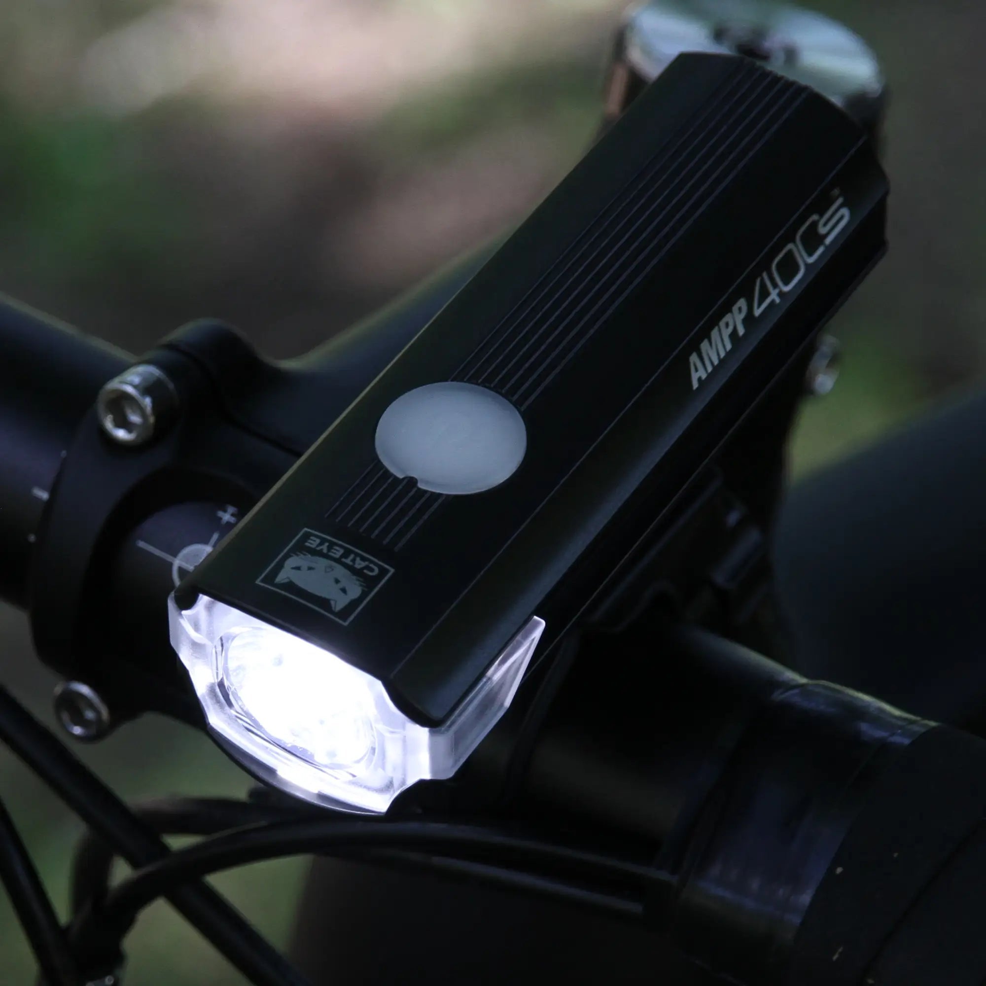 Close-up of the CATEYE AMPP400S black bike light mounted on handlebars, shining a bright white beam in daylight, with its USB-C charging port visible.