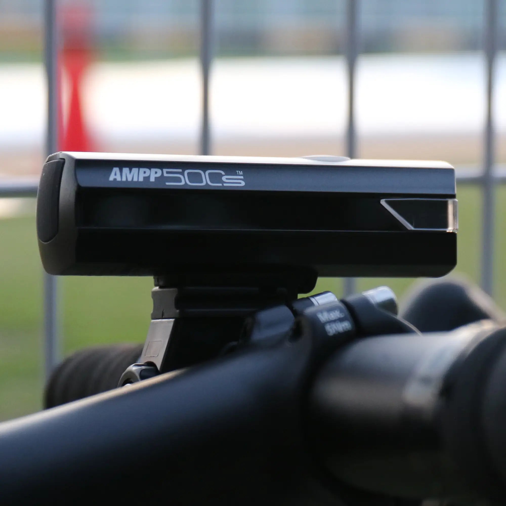 A close-up of a black CATEYE AMPP500S bicycle front light with OptiCube™ lens, mounted securely on handlebars using a FlexTight™ bracket, with a blurred fence and outdoor background.